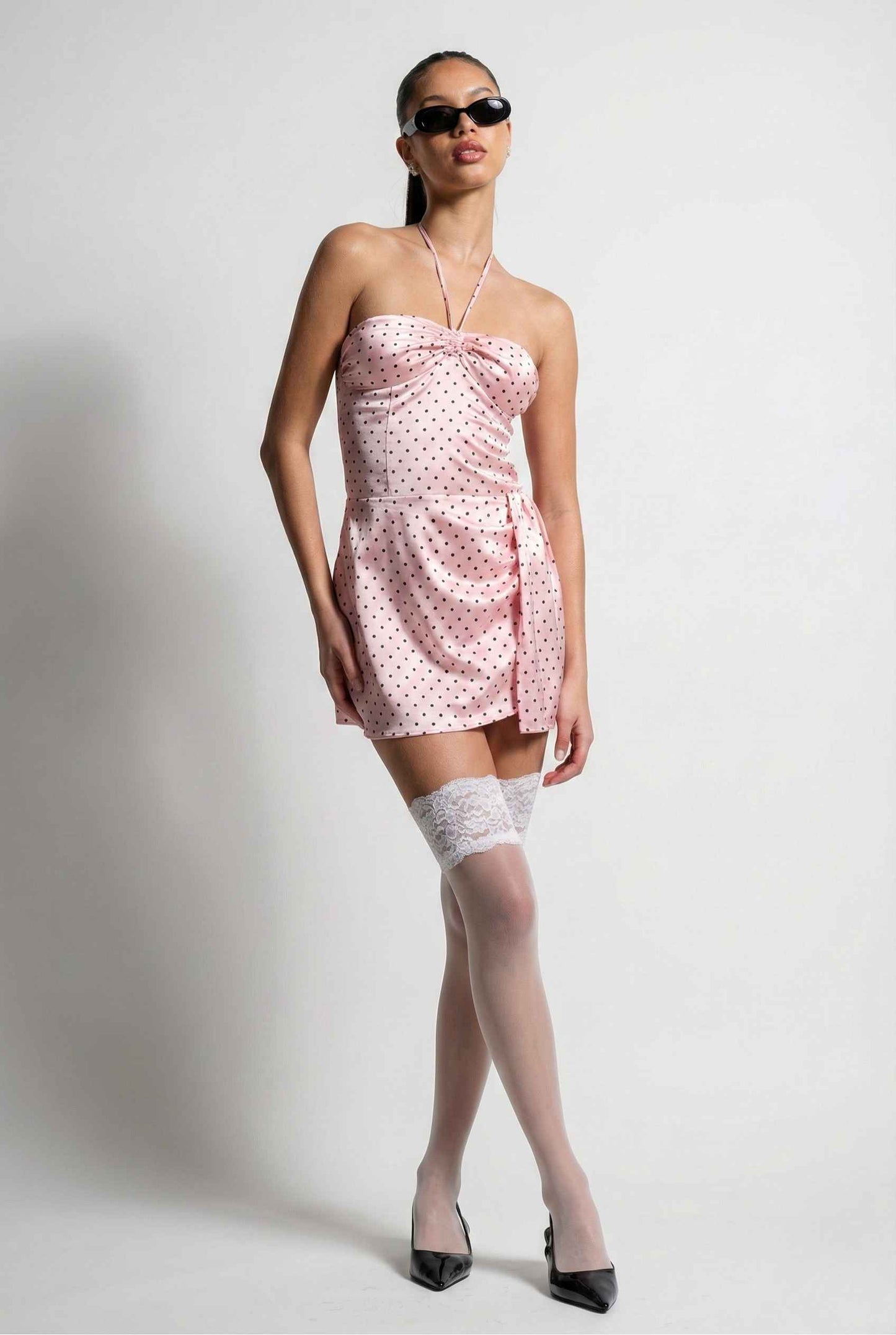 Woman wearing a Lola Mini Pink Polka Dot Satin Halter Dress with white stockings and black sunglasses on a plain background
