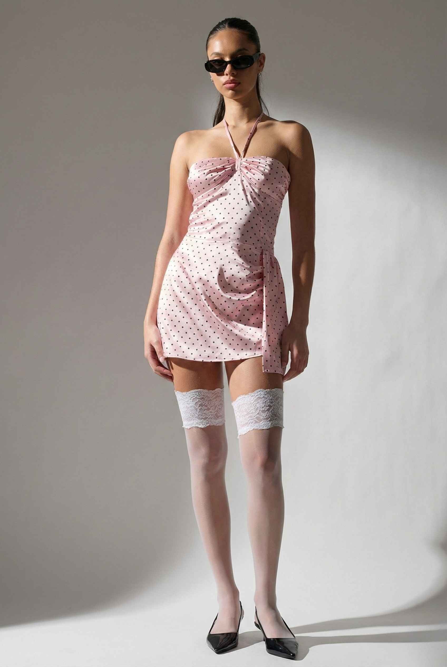 Model wearing a Lola Mini Pink Polka Dot Satin Halter Dress with white polka dots and white knee-high socks against a plain background.