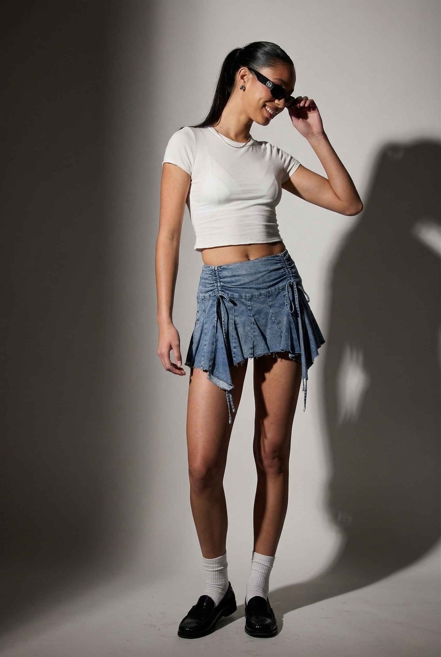 Model wearing Lola Skirt in light Blue Denim Ruched Waist Mini with Ruffle Hem with basic cropped t-shirt, socks and loafers