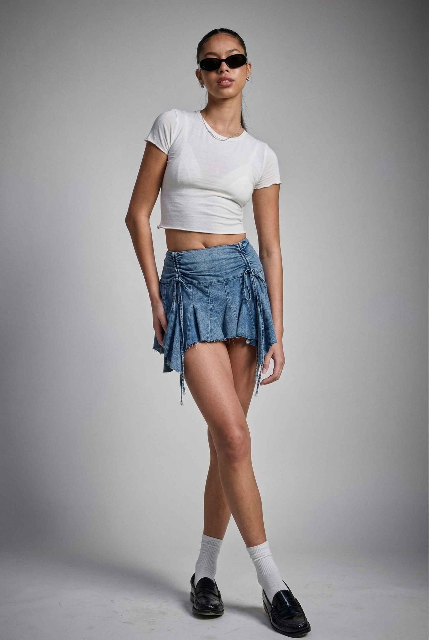Model wearing Lola Skirt in light Blue Denim Ruched Waist Mini with Ruffle Hem with basic cropped t-shirt, socks and loafers