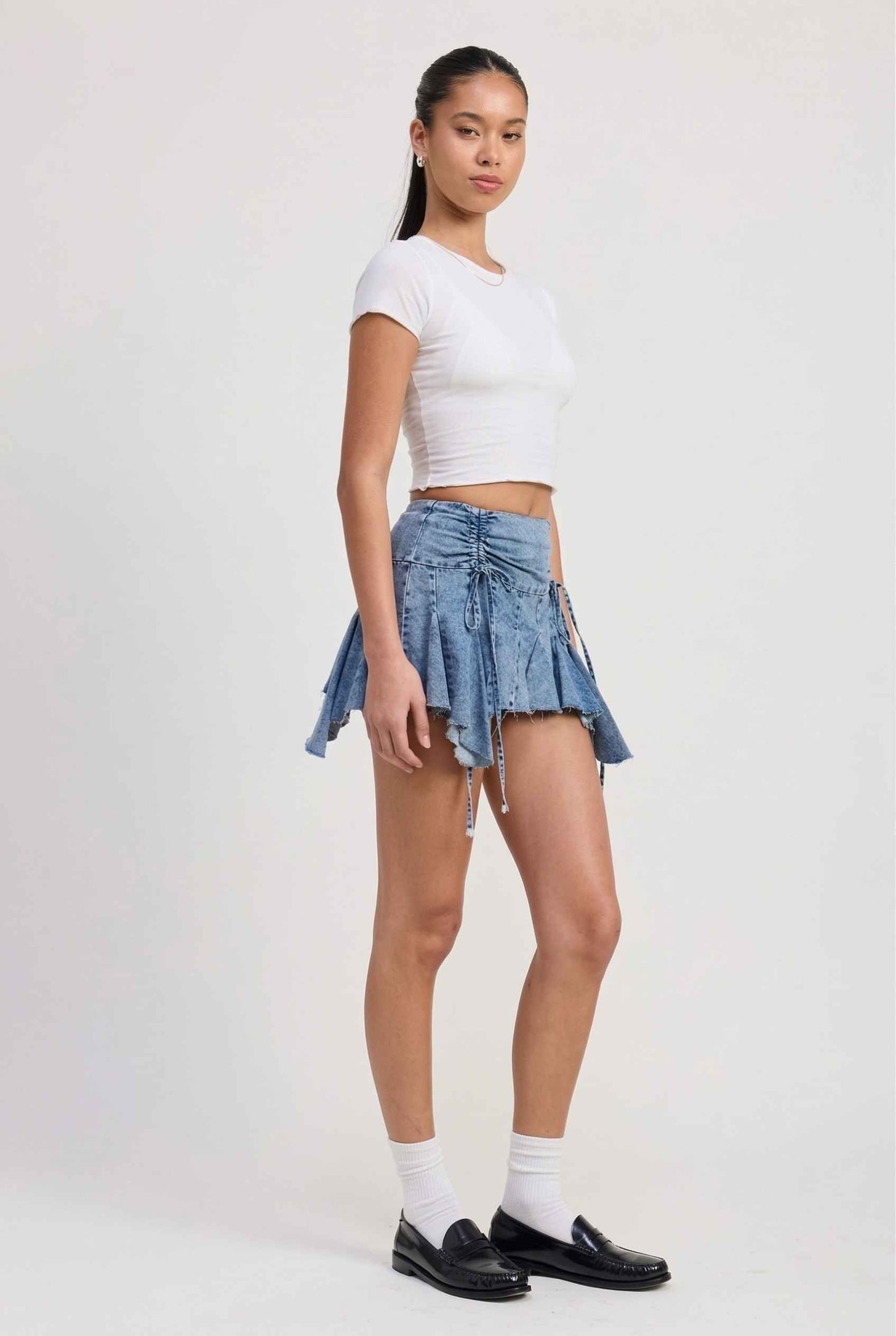 Model wearing Lola Skirt in light Blue Denim Ruched Waist Mini with Ruffle Hem with basic cropped t-shirt, socks and loafers