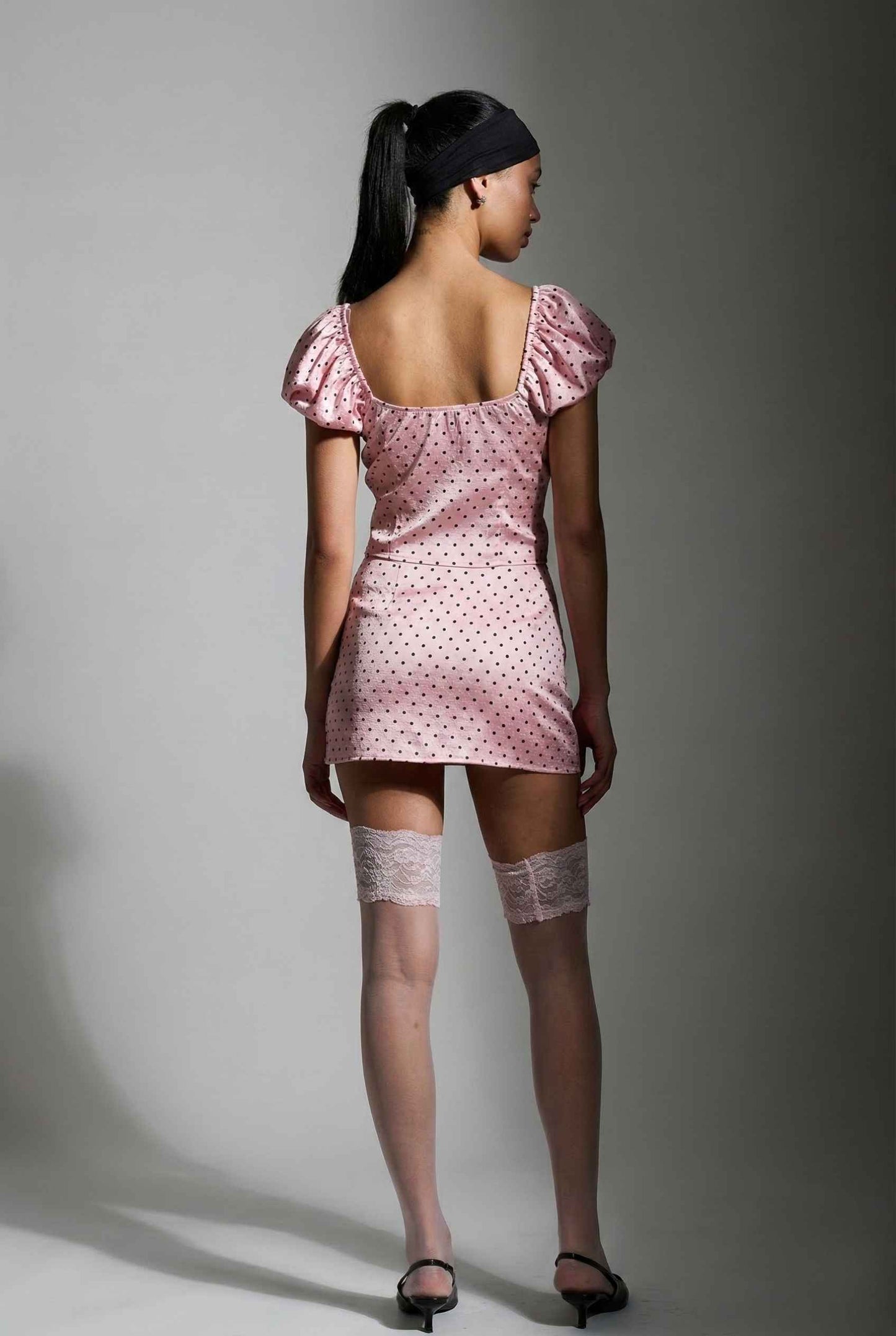 Woman wearing a Lola Top Pink & Black Polka Dot Puff Sleeve Tie-Front Blouse with puffed sleeves and lace stockings against a gray background