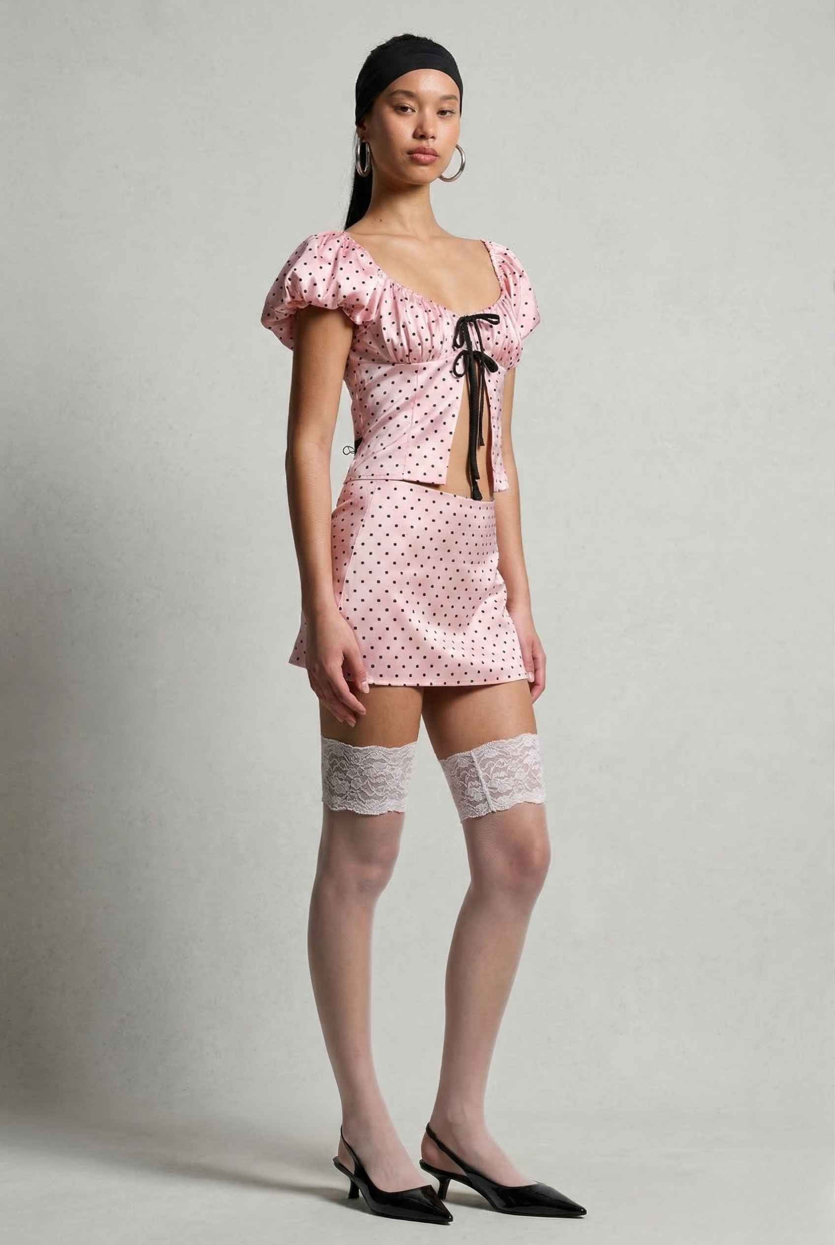 Woman wearing a Lola Top Pink & Black Polka Dot Puff Sleeve Tie-Front Blouse with a black belt on a gray background