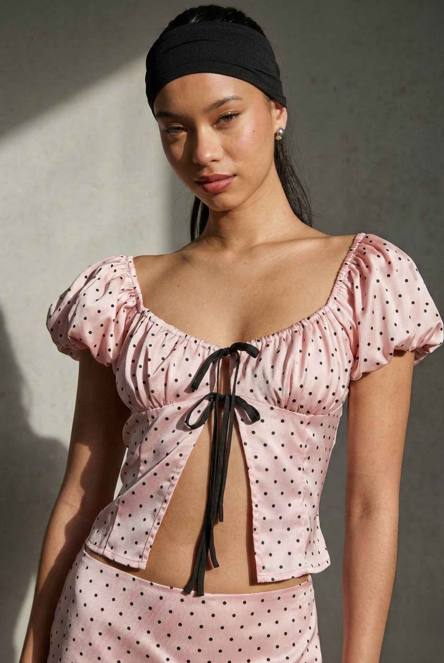 Woman wearing a Lola Top Pink & Black Polka Dot Puff Sleeve Tie-Front Blouse with puff sleeves against a gray background
