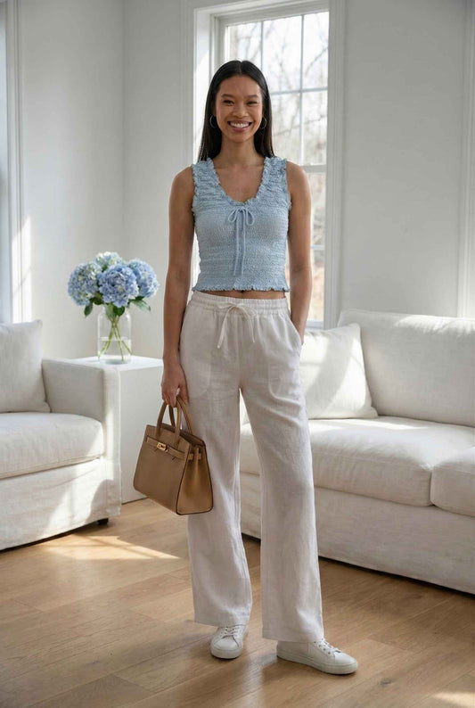 Woman in Luna Top Light Blue Smocked Ruffle Tie-Front Tank and white pants standing in a bright living room.