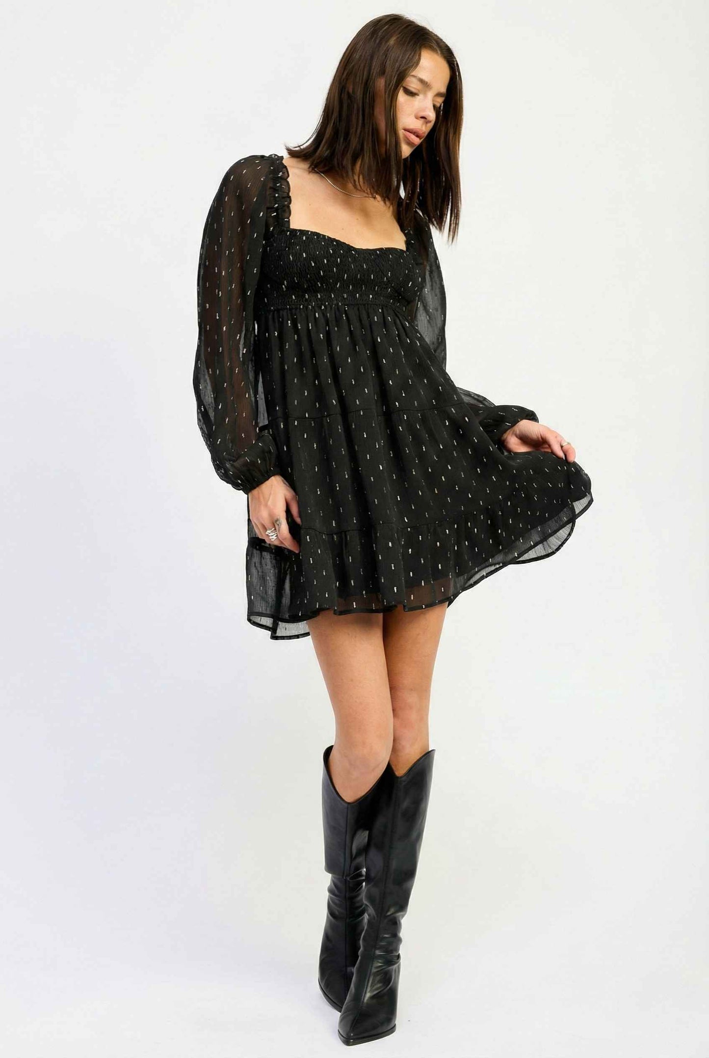 Woman wearing a Lyra Mini Black Smocked Long Sleeve Dress with sheer sleeves and a white background