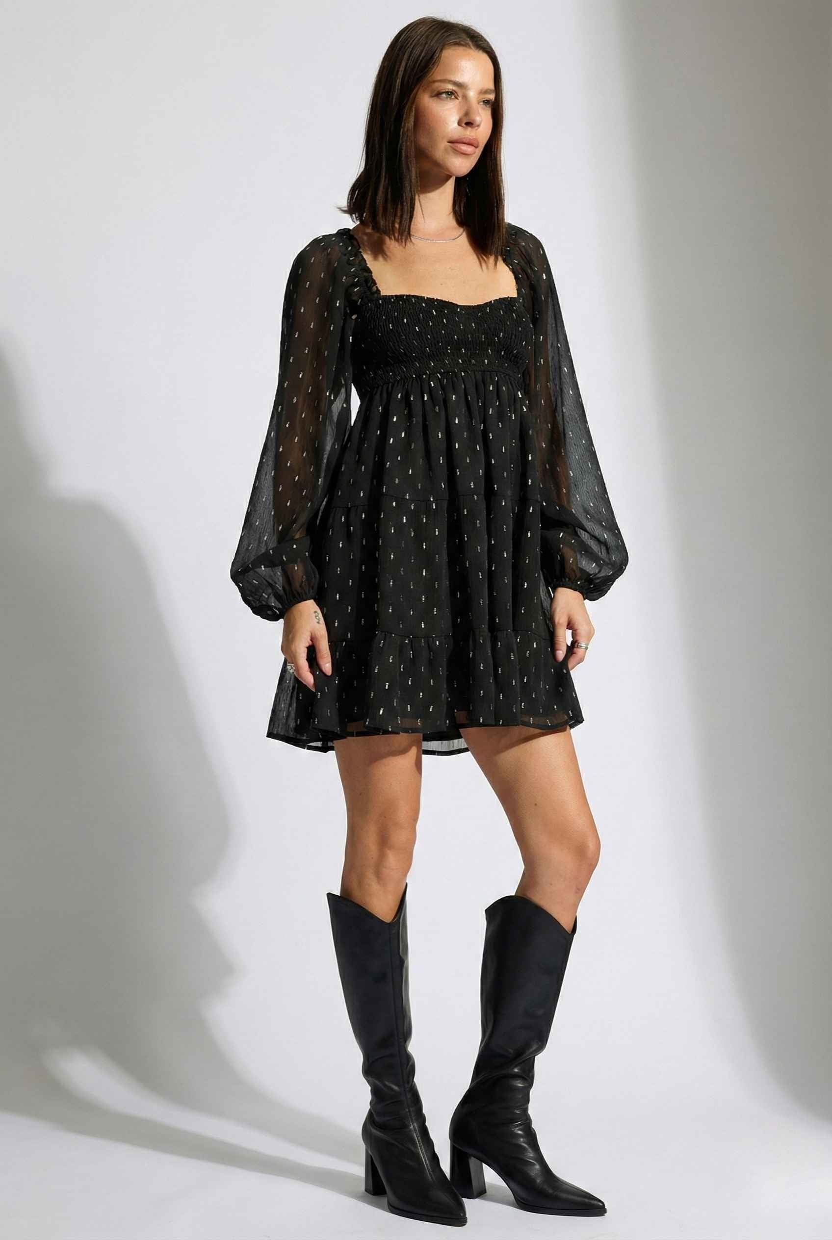 Woman wearing a Lyra Mini Black Smocked Long Sleeve Dress with sheer sleeves and knee-high boots on a white background