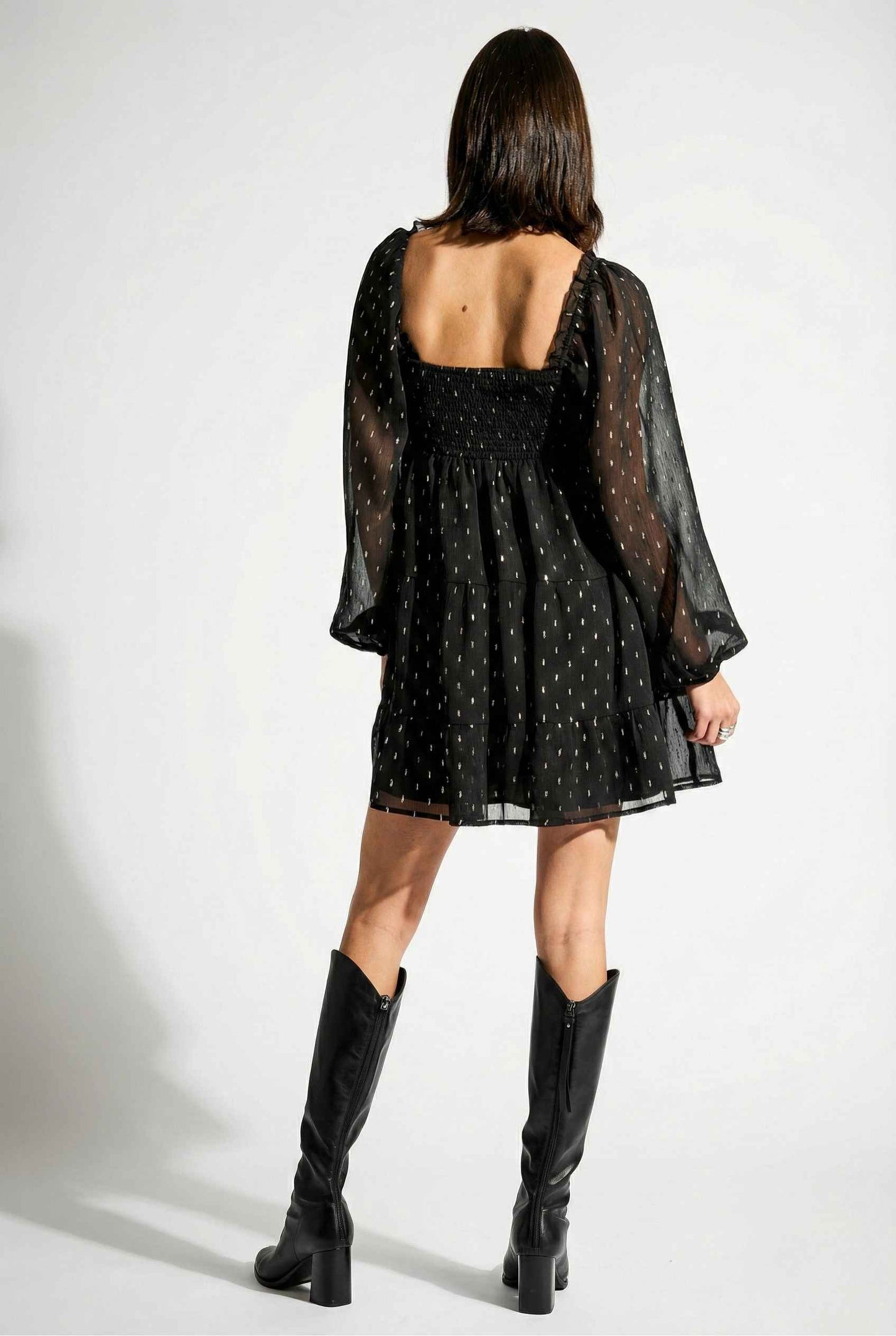 Woman wearing a Lyra Mini Black Smocked Long Sleeve Dress with sheer sleeves and black knee-high boots on a white background