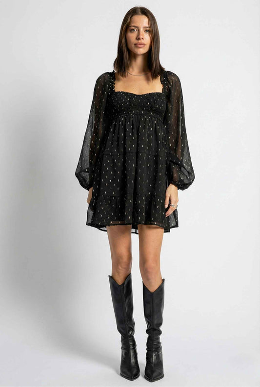 Woman wearing a Lyra Mini Black Smocked Long Sleeve Dress with sheer sleeves and black boots on a white background