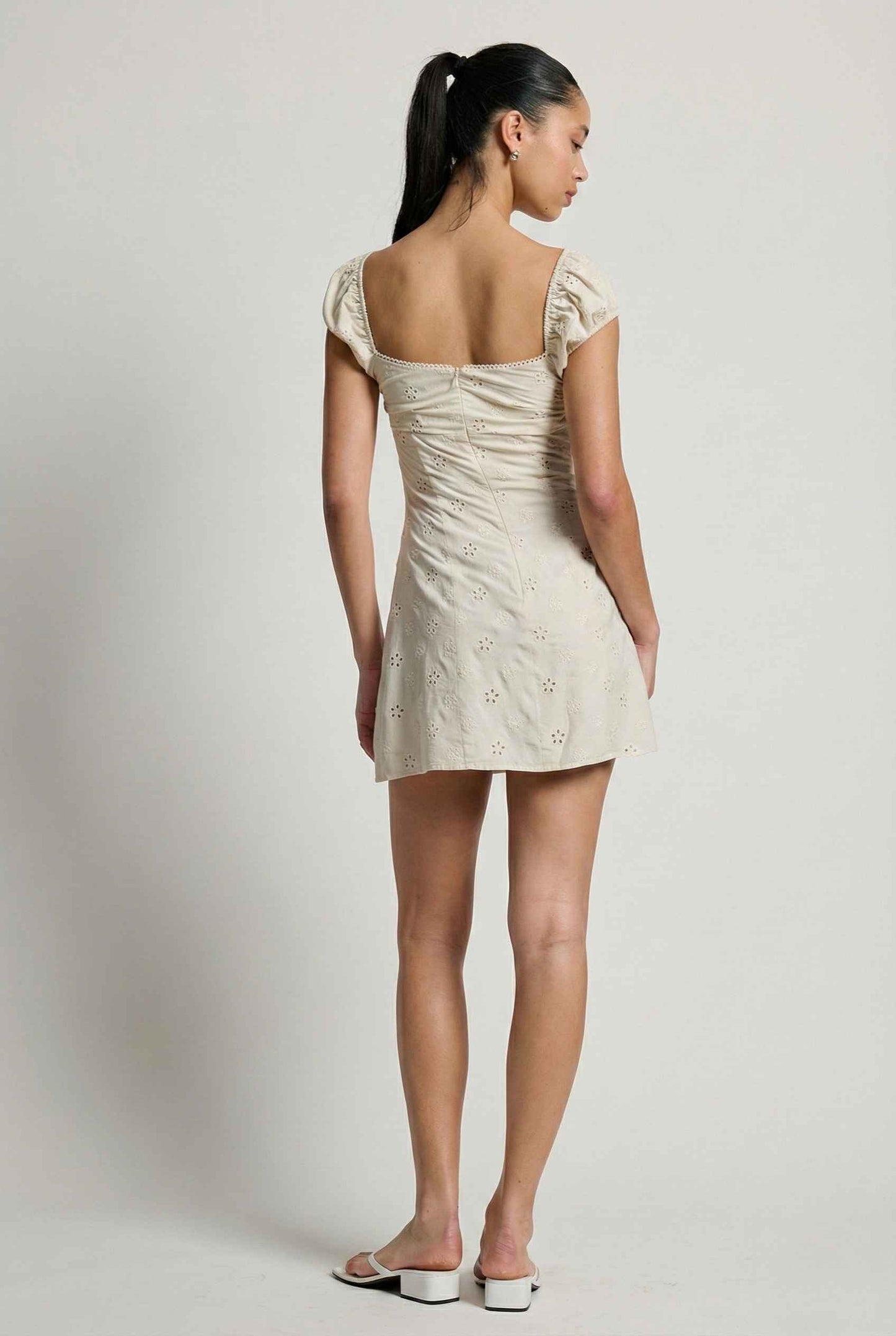 Woman wearing a Maeve Mini Ivory Eyelet Ruched Puff Sleeve Dress with cap sleeves on a plain background