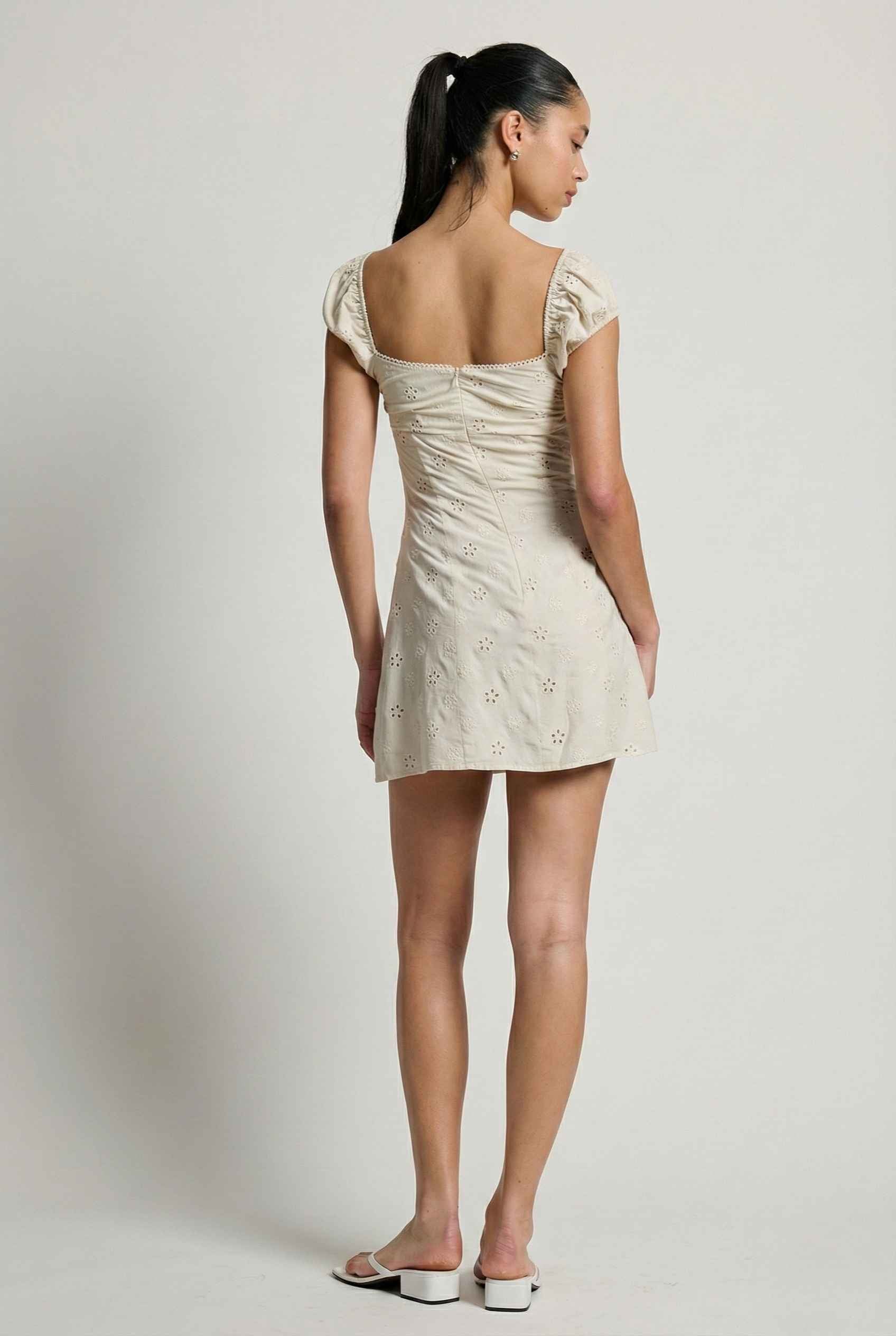 Woman wearing a Maeve Mini Ivory Eyelet Ruched Puff Sleeve Dress with cap sleeves on a plain background