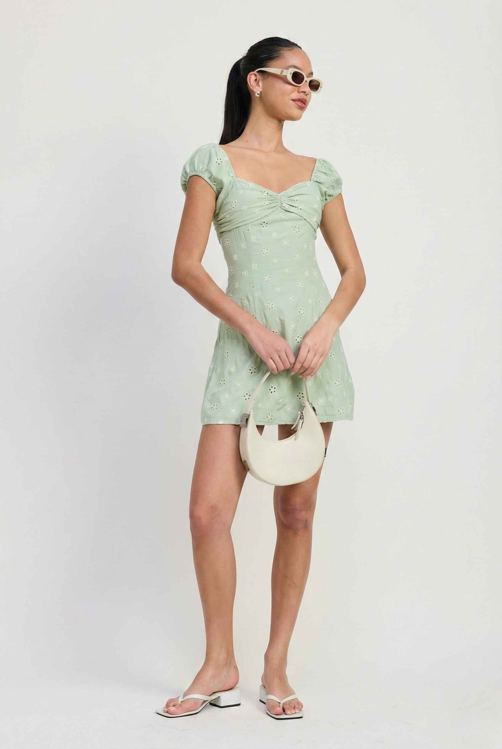 Woman wearing a Maeve Mini Mint Eyelet Ruched Puff Sleeve Dress holding a white handbag against a plain background