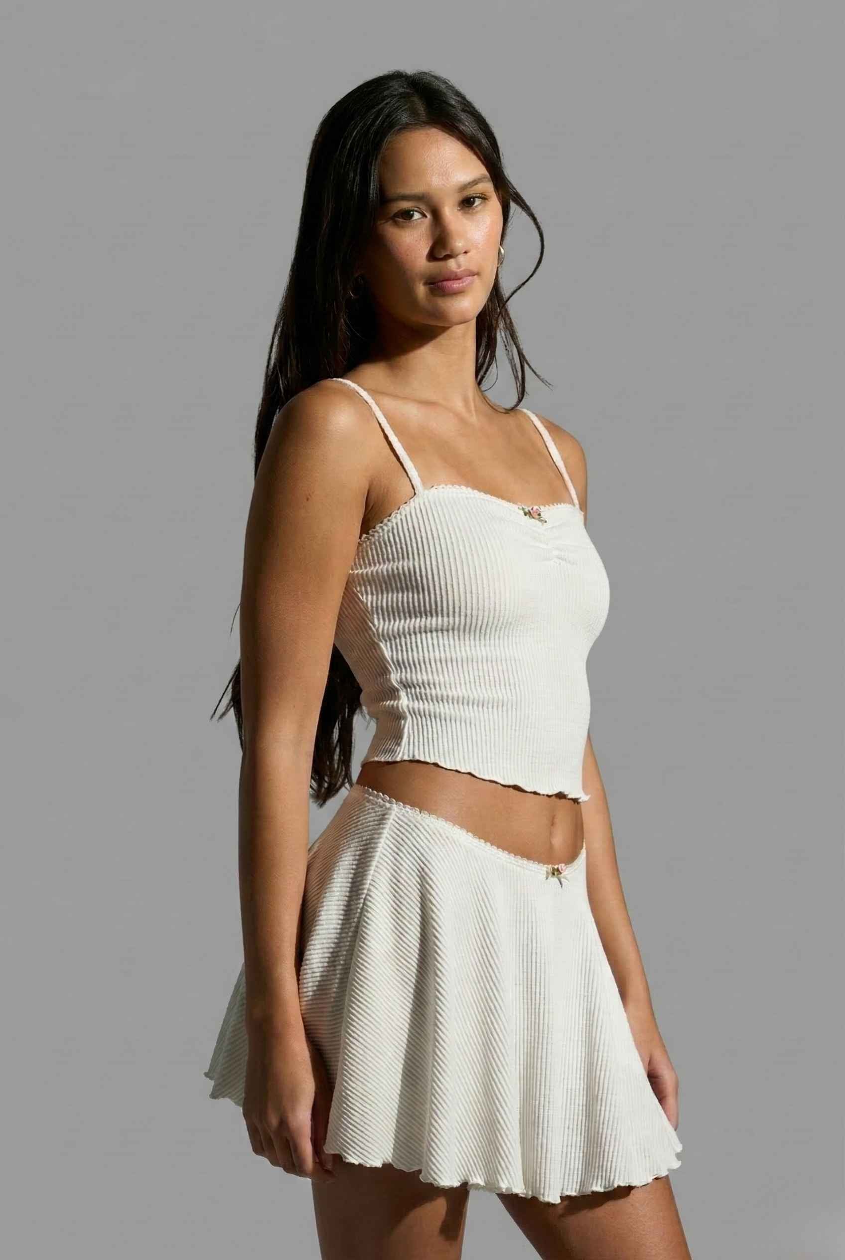 Woman wearing a Maisie Cami White Ribbed Knit Bow Top with Picot Trim and skirt against a gray background