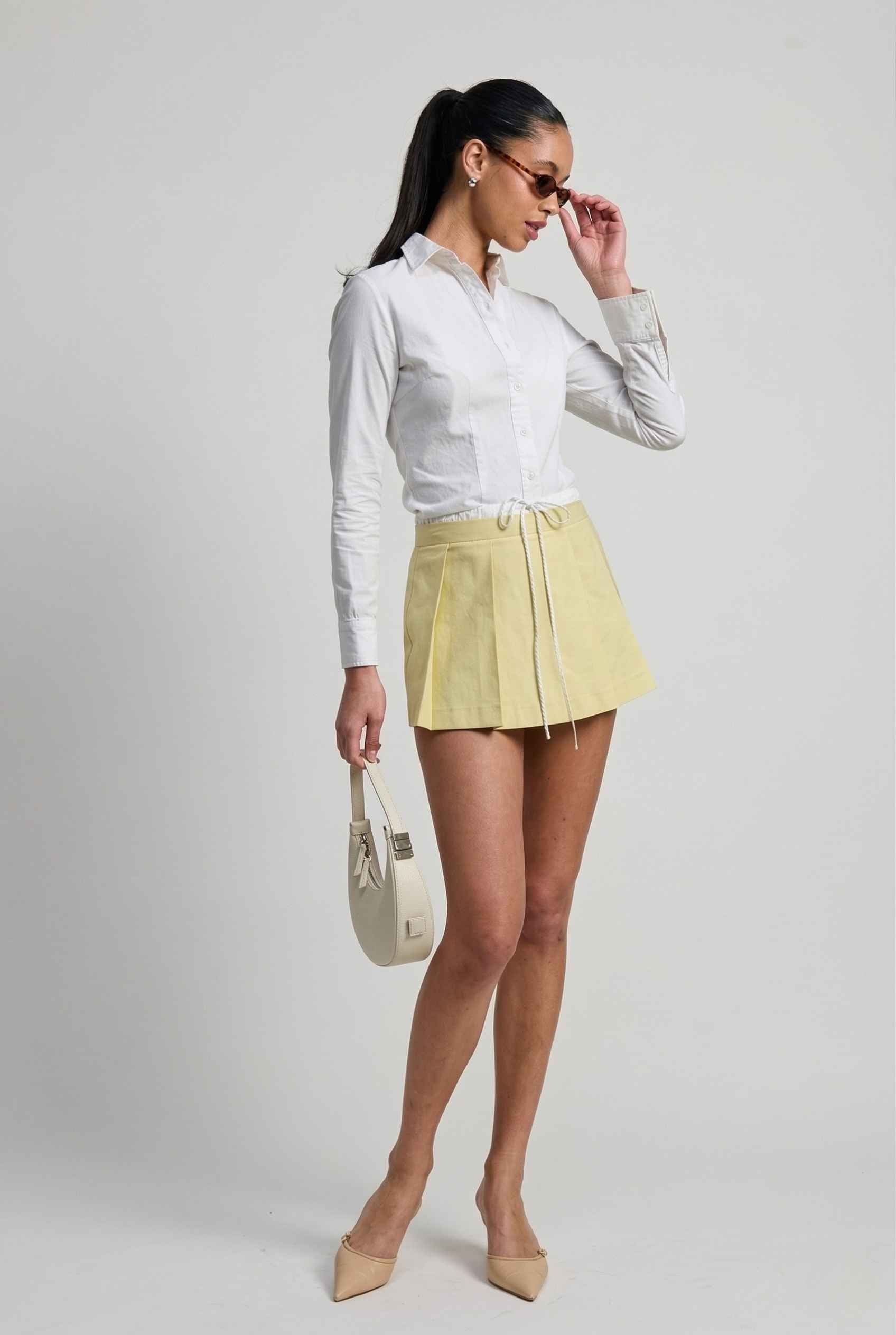 Model wearing Margo Skort in Yellow Poplin Cotton Twill Pleated Mini Skort paired with crisp poplin blouse and basic heels