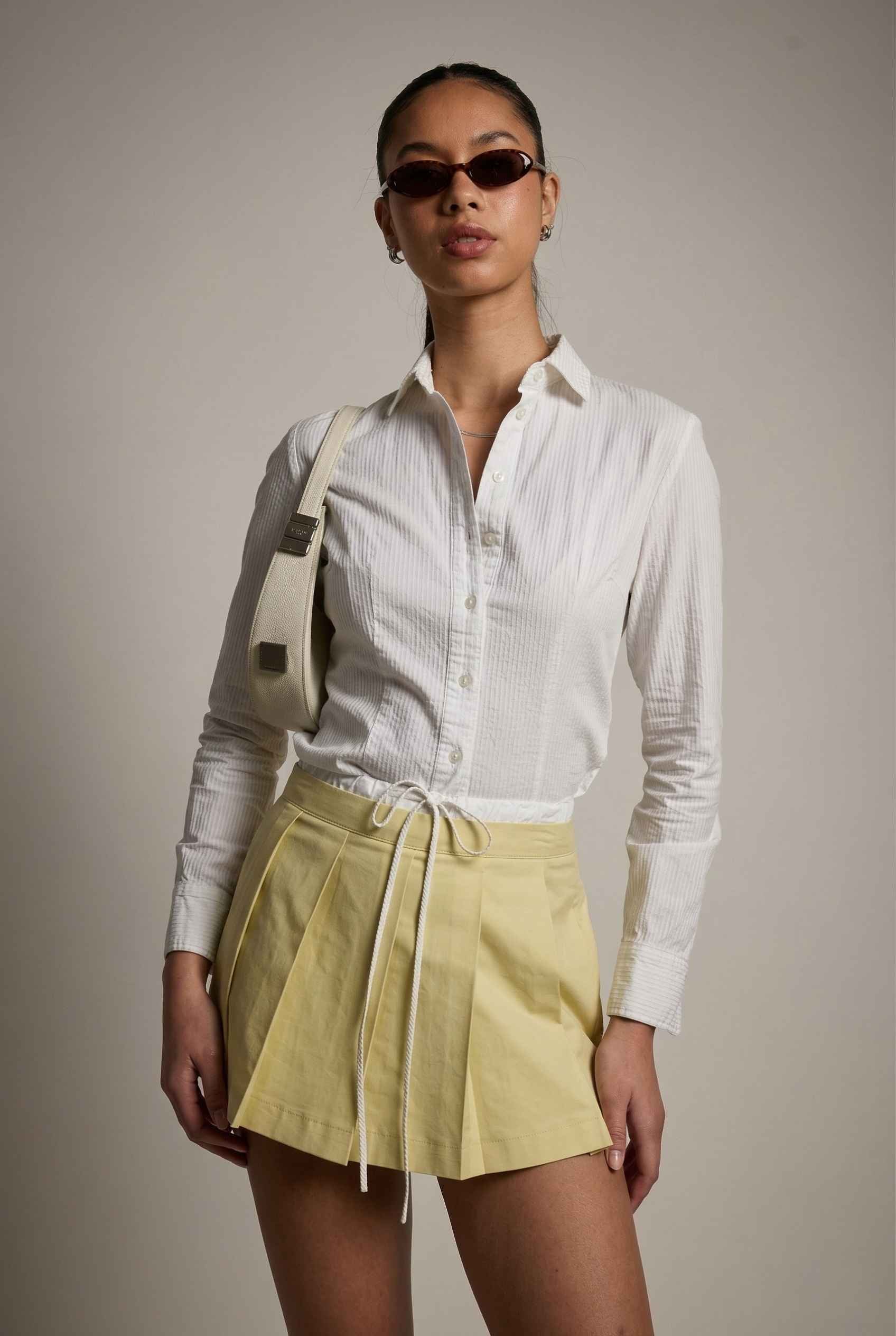 Model wearing Margo Skort in Yellow Poplin Cotton Twill Pleated Mini Skort paired with crisp poplin blouse and handbag