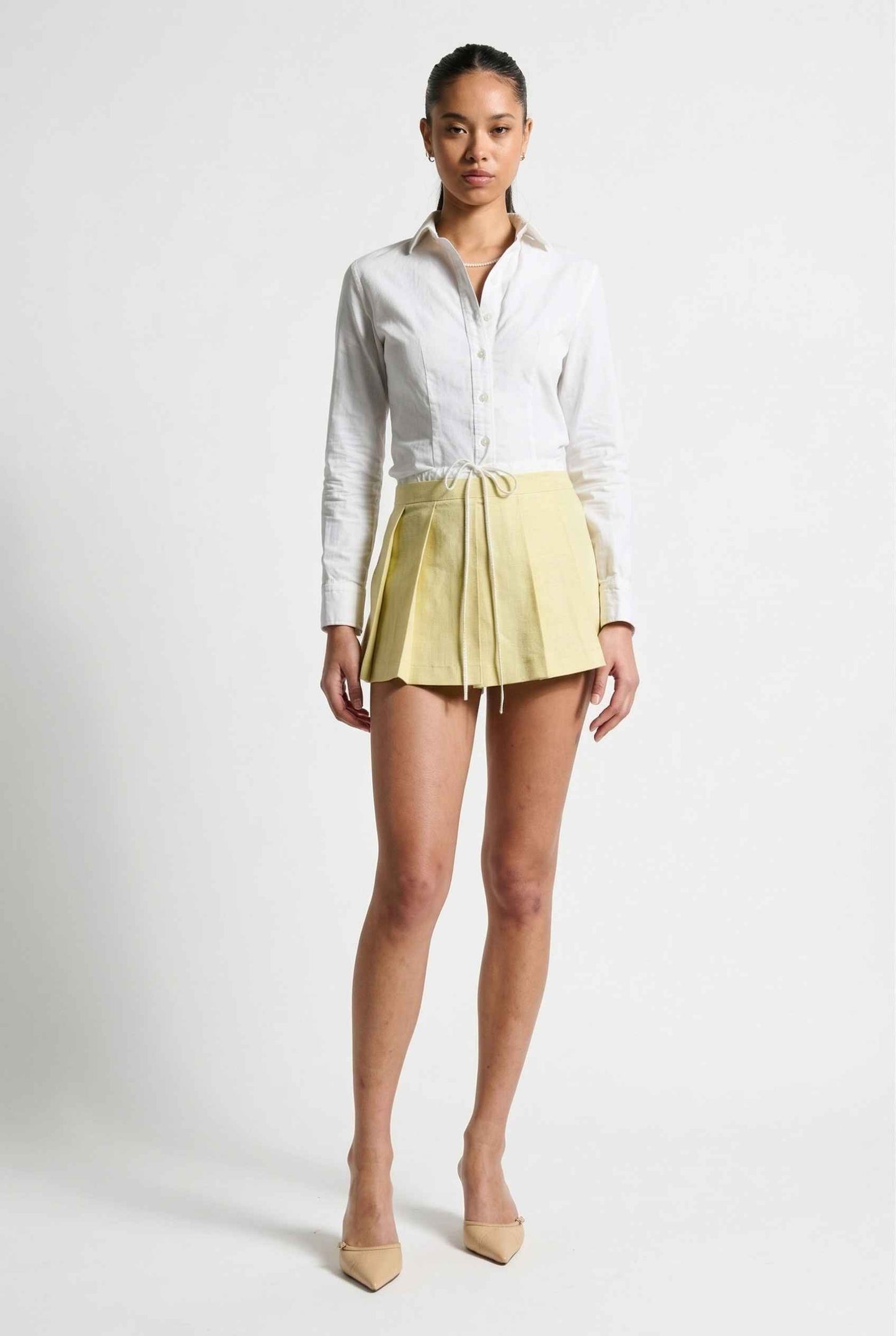 Model wearing Margo Skort in Yellow Poplin Cotton Twill Pleated Mini Skort paired with crisp poplin blouse and basic heels