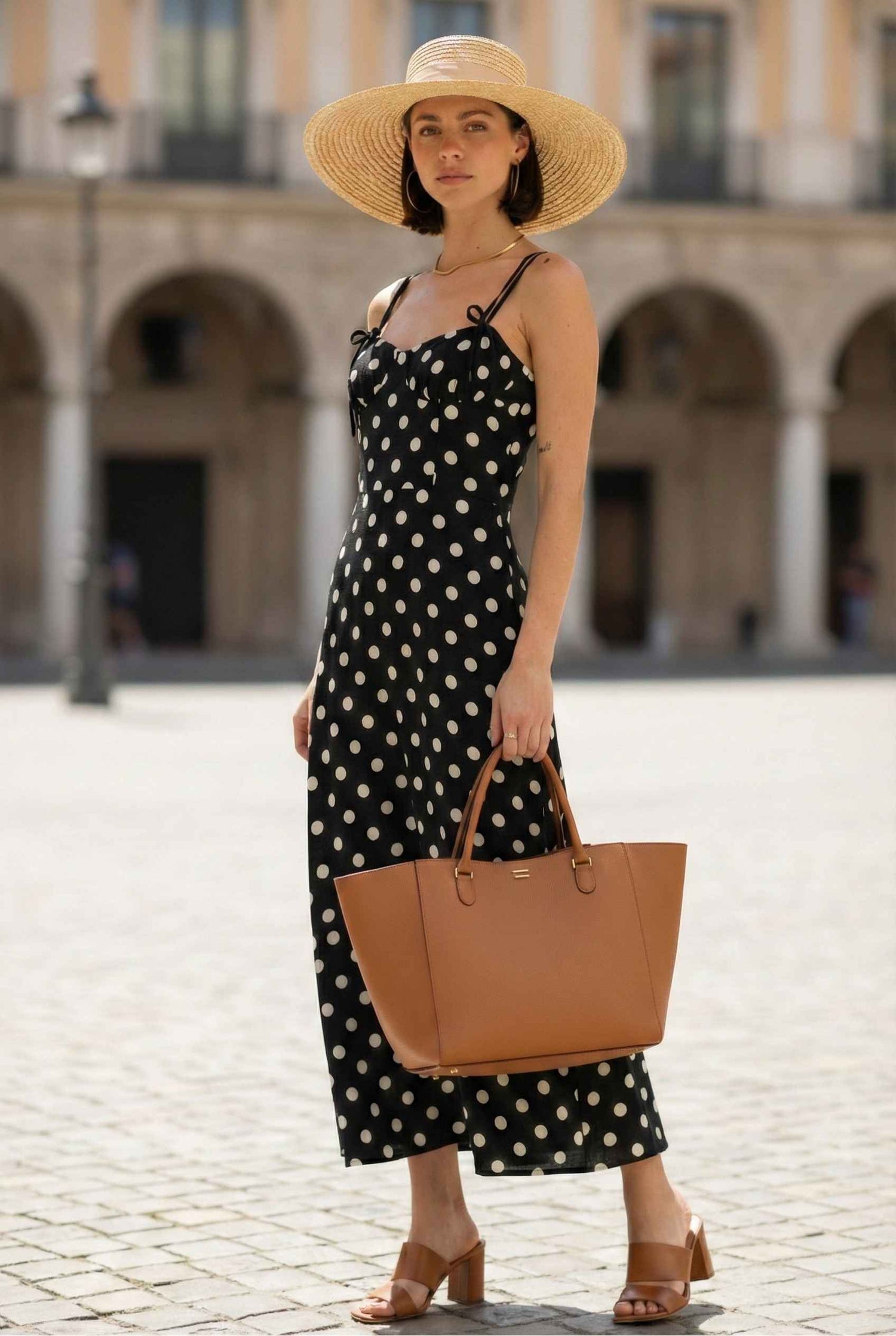 Woman in a Margot Maxi Black Polka Dot Linen Blend Bustier Dress and straw hat holding a brown bag in an outdoor setting.
