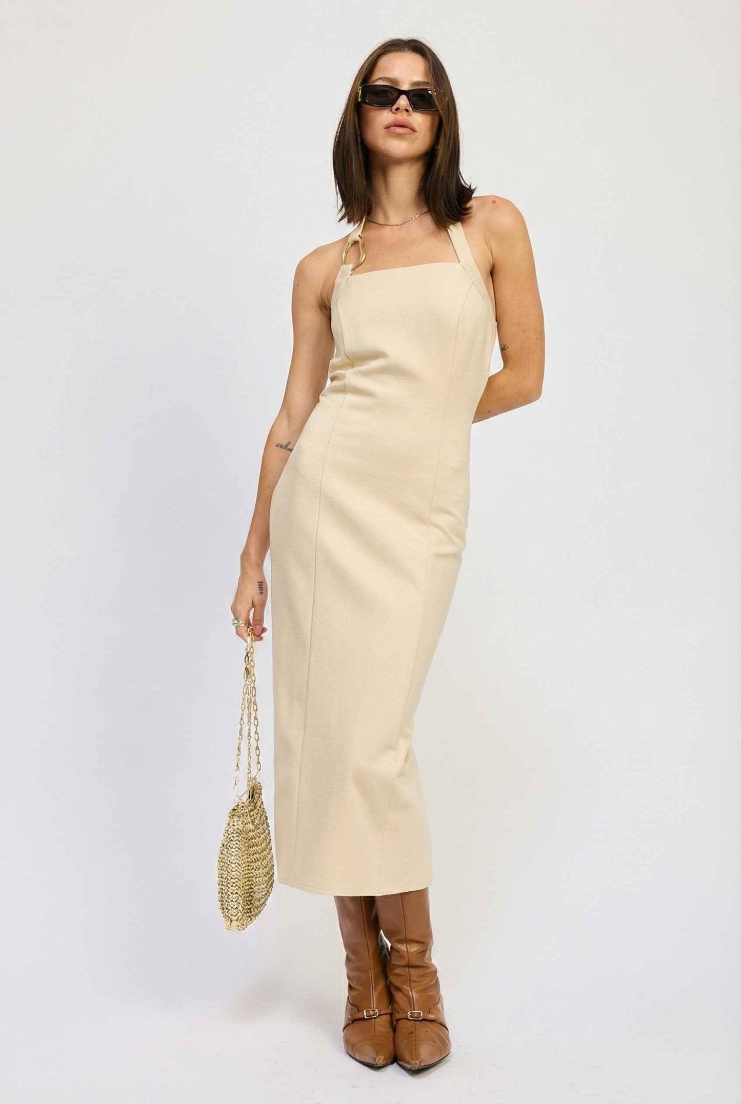 Woman wearing a Mercer Midi Taupe Halter Hardware Dress with brown boots and a straw bag on a white background