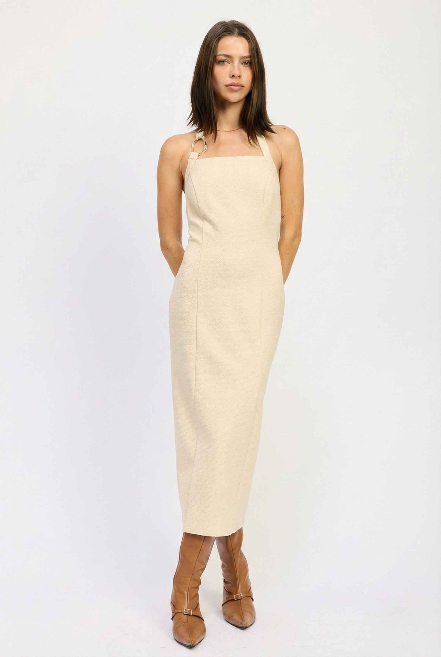 Woman wearing a Mercer Midi Taupe Halter Hardware Dress with brown boots on a white background