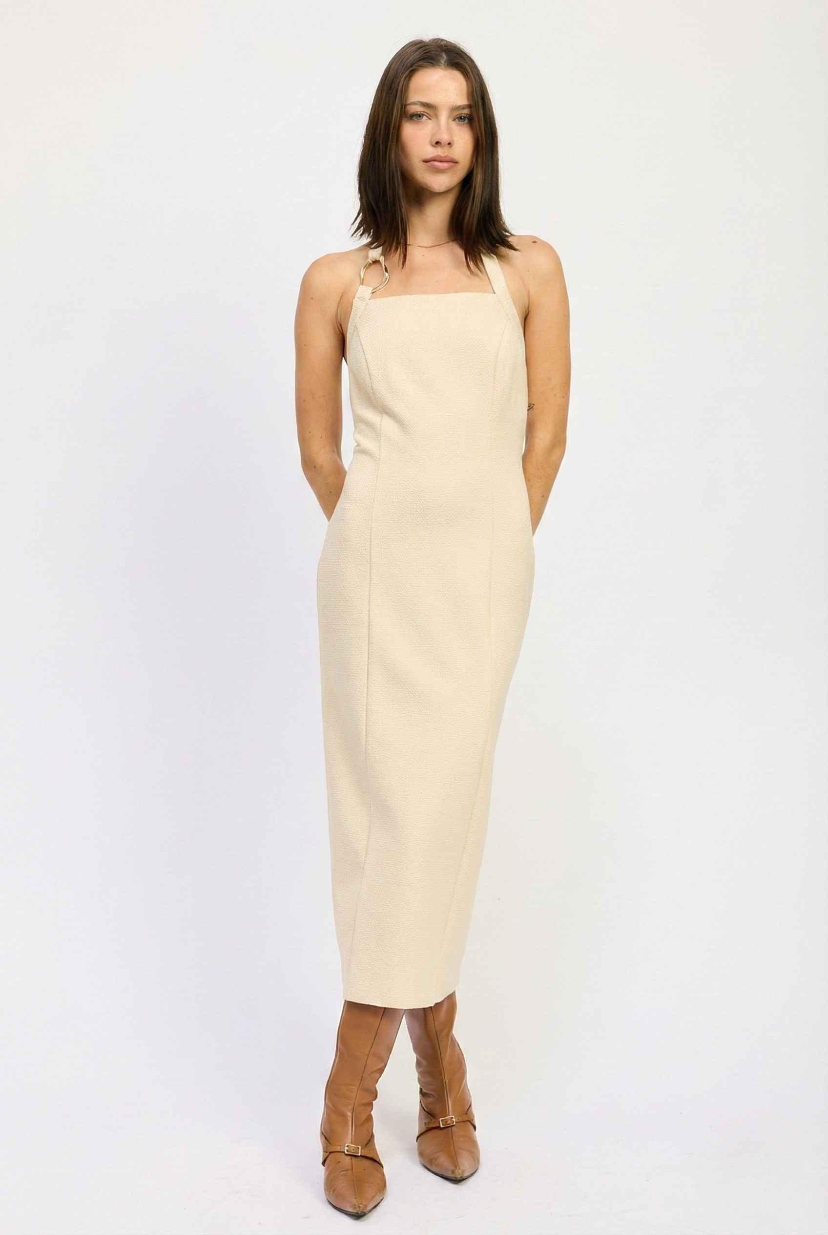 Woman wearing a Mercer Midi Taupe Halter Hardware Dress with brown boots on a white background