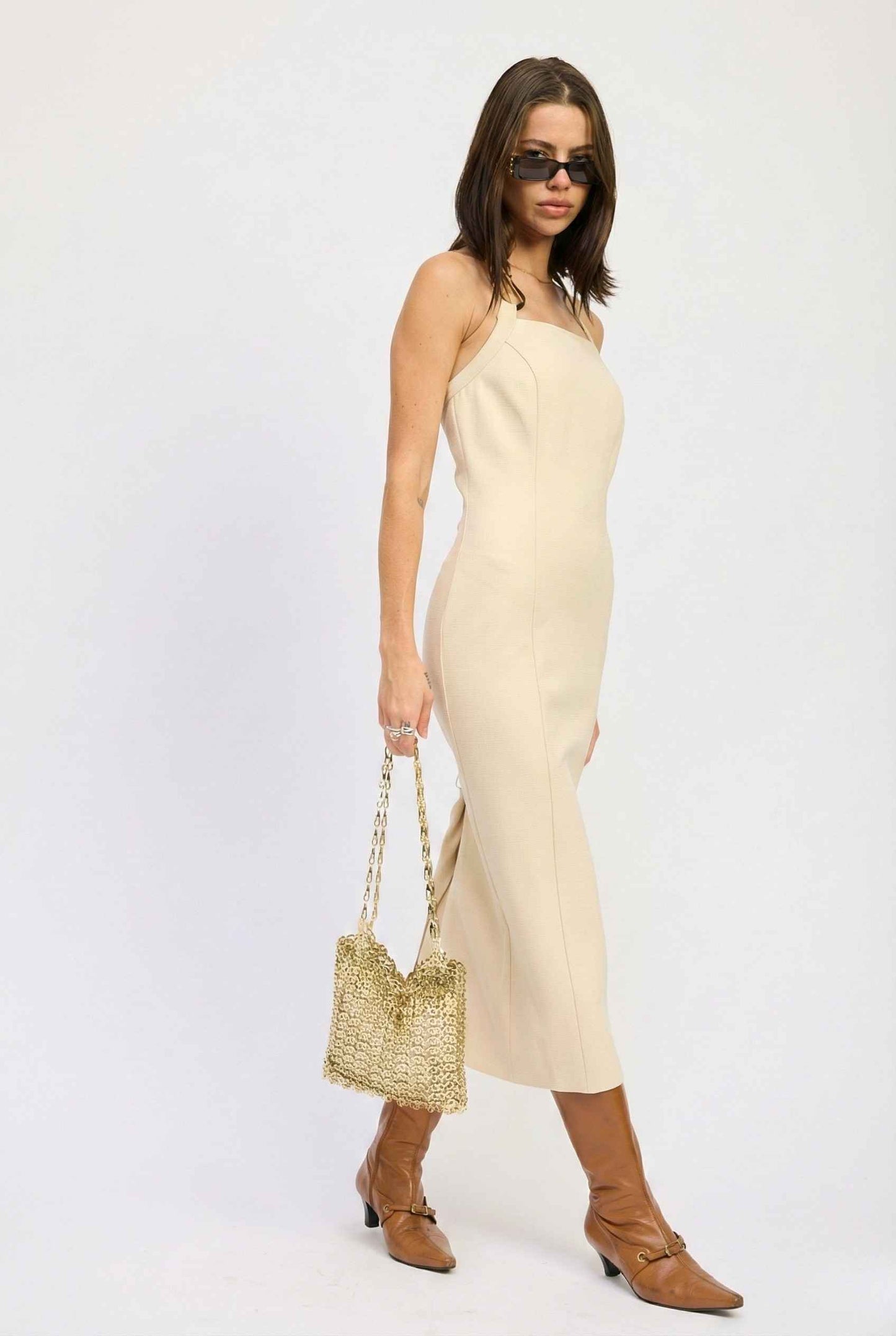 Woman wearing a Mercer Midi Taupe Halter Hardware Dress with a straw bag on a white background