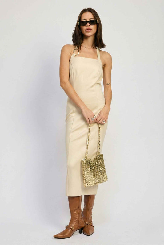 Woman wearing a Mercer Midi Taupe Halter Hardware Dress and brown boots, holding a gold bag against a white background