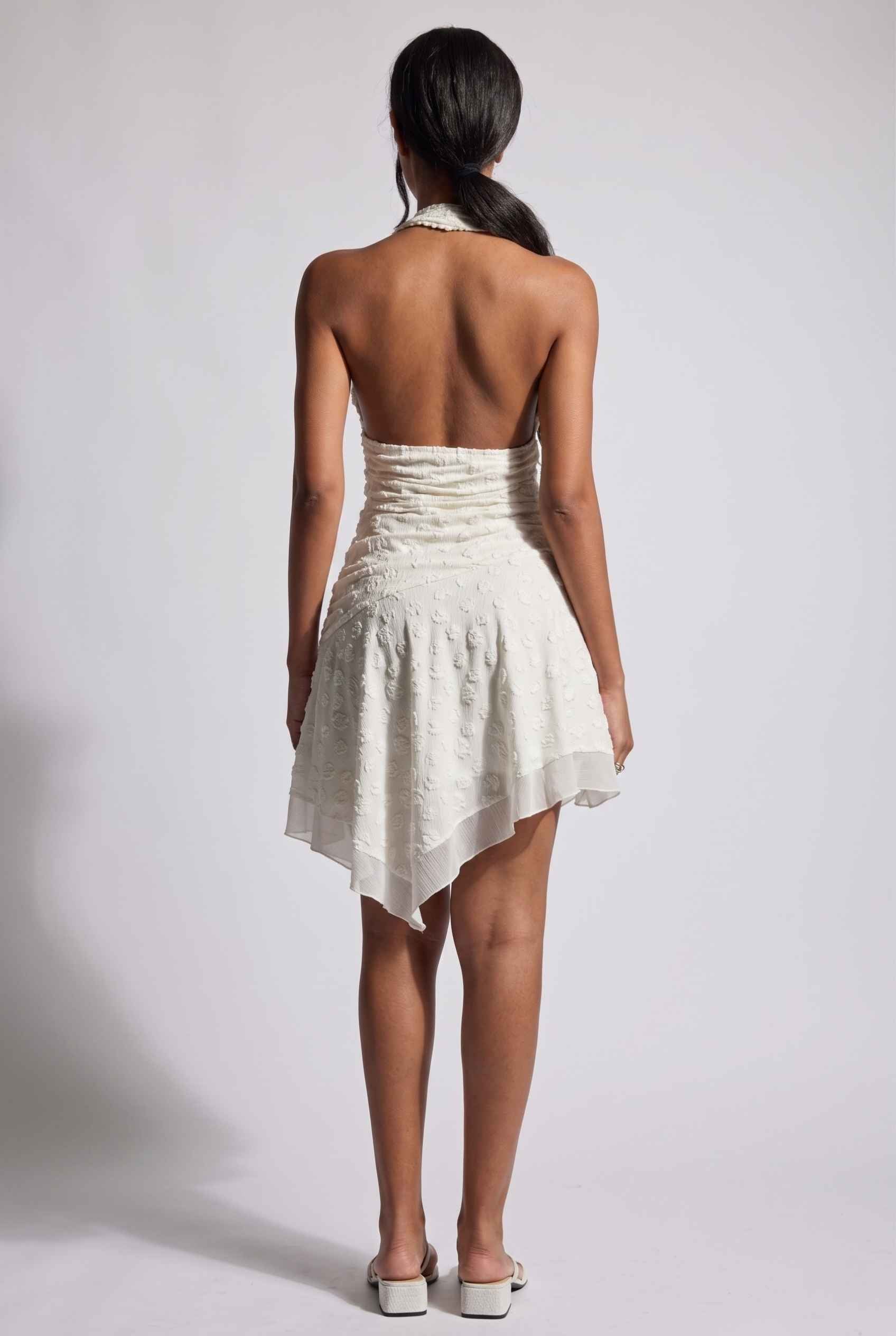 Model wearing Mikala Ruched Halter Mini Dress in Ivory Cocktail Dress paired with minimalist heels back view open back