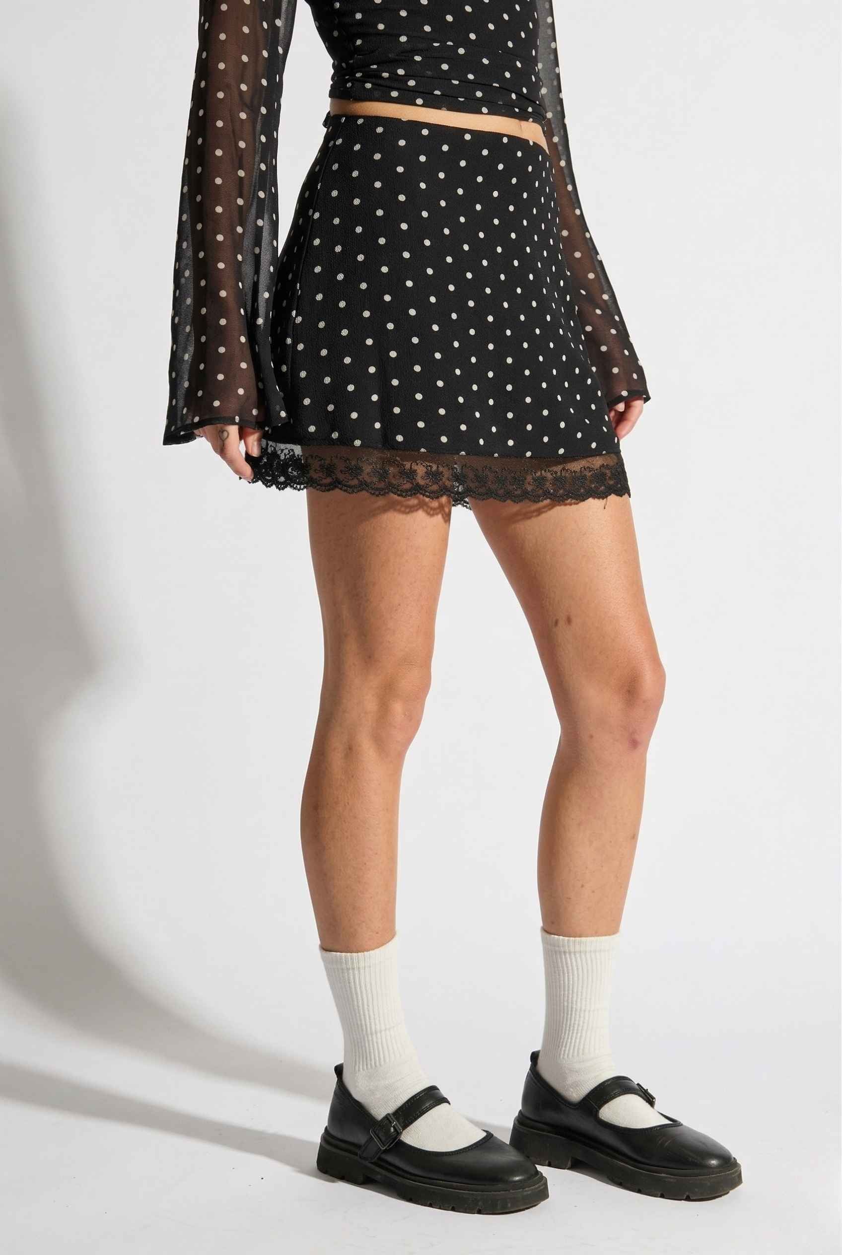 Woman wearing a black polka dot top and Mila Skirt Black Polka Dot Mesh Mini Skirt with Lace Trim with white socks and black shoes on a white background