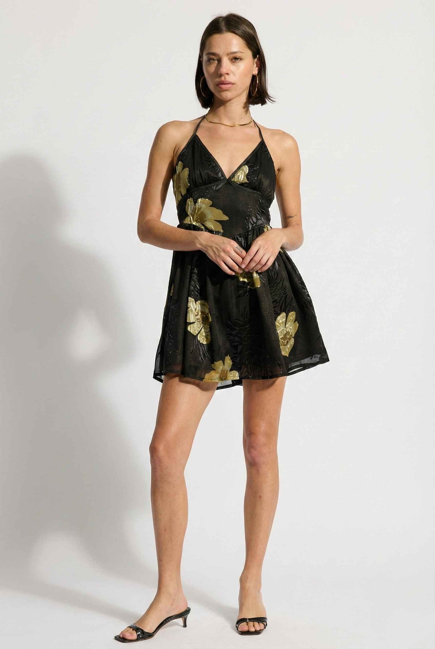 Woman wearing a Nina Mini Black & Gold Floral Halter Dress with gold floral patterns on a white background