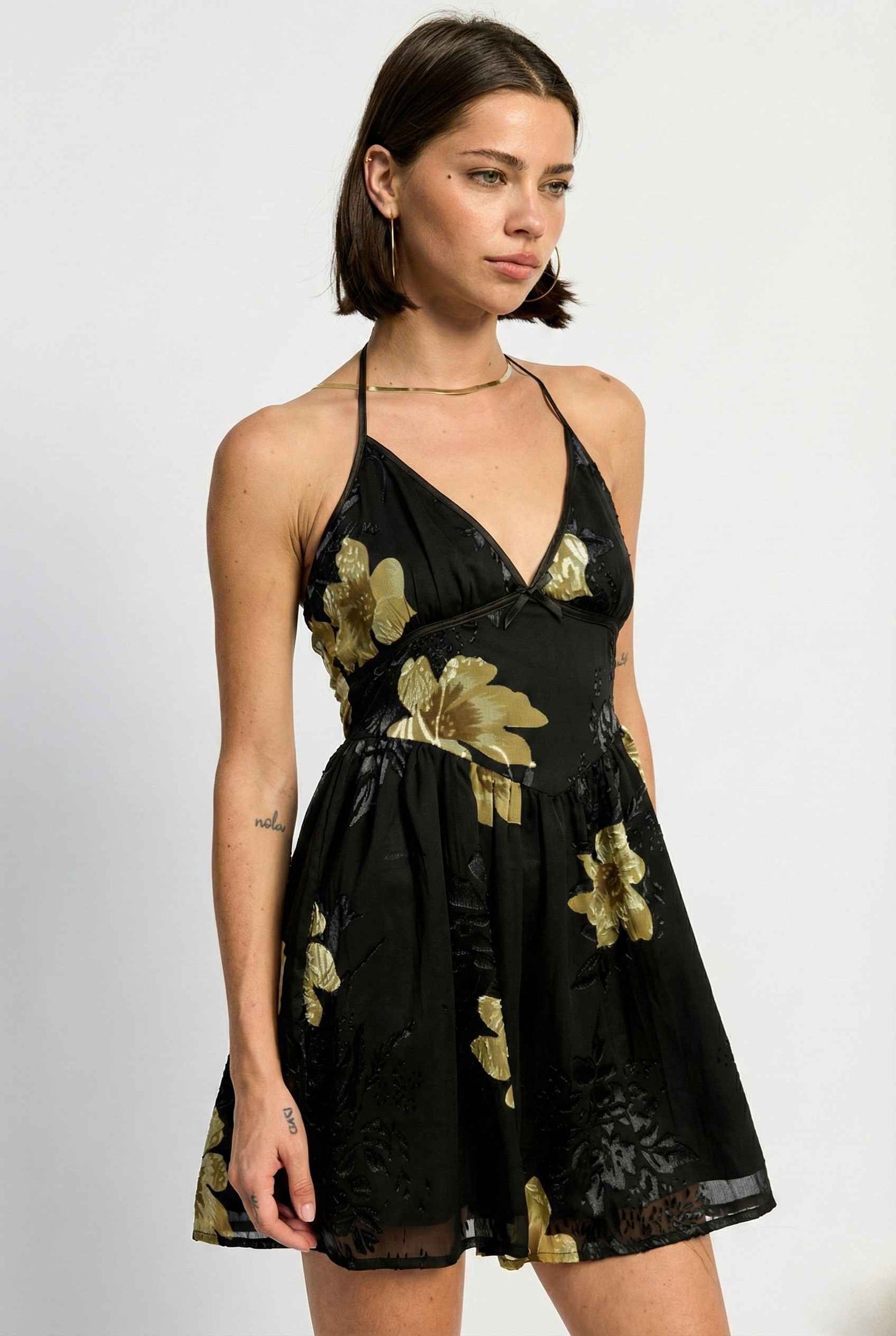Woman wearing a Nina Mini Black & Gold Floral Halter Dress with gold floral patterns on a white background
