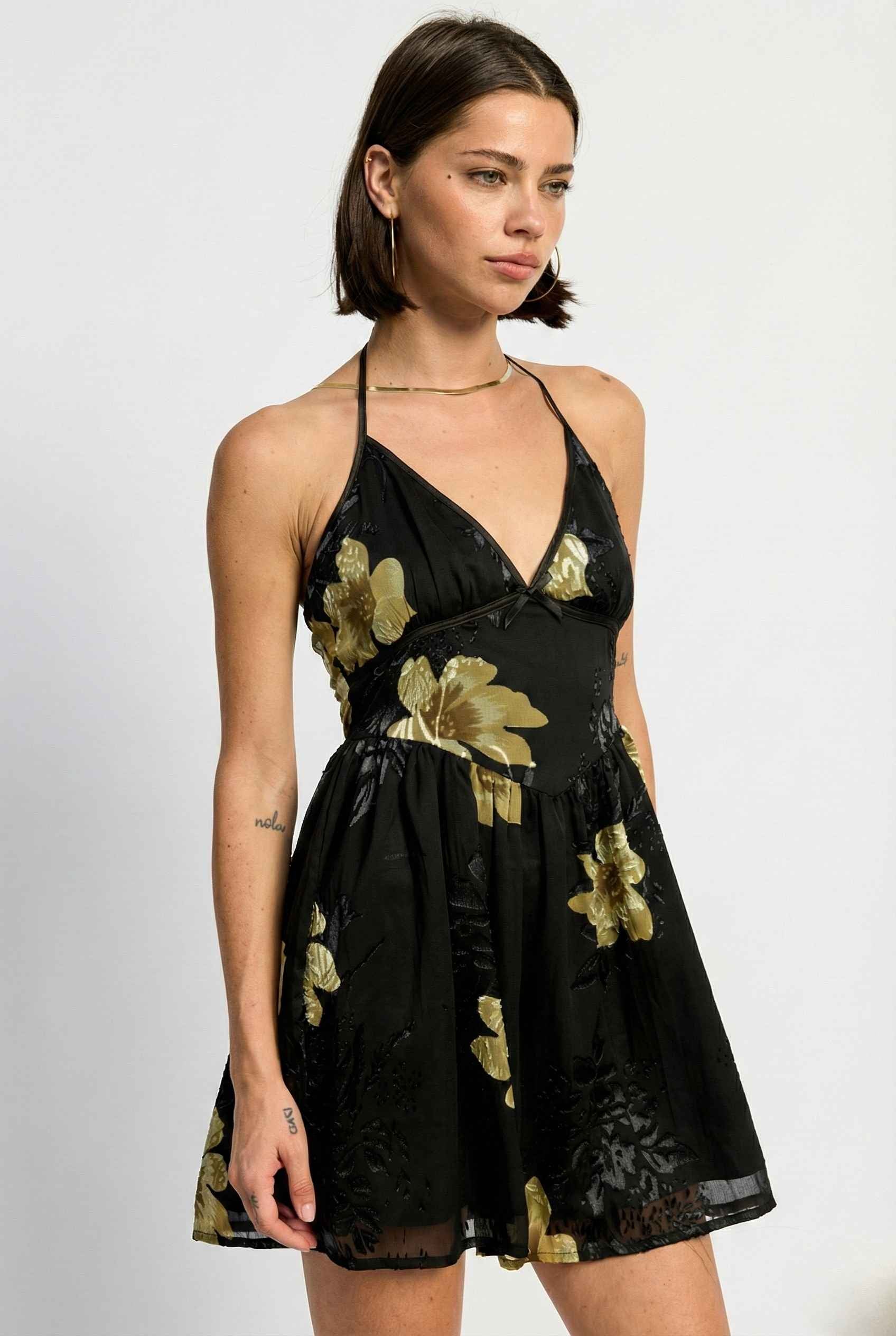 Woman wearing a Nina Mini Black & Gold Floral Halter Dress with gold floral patterns on a white background
