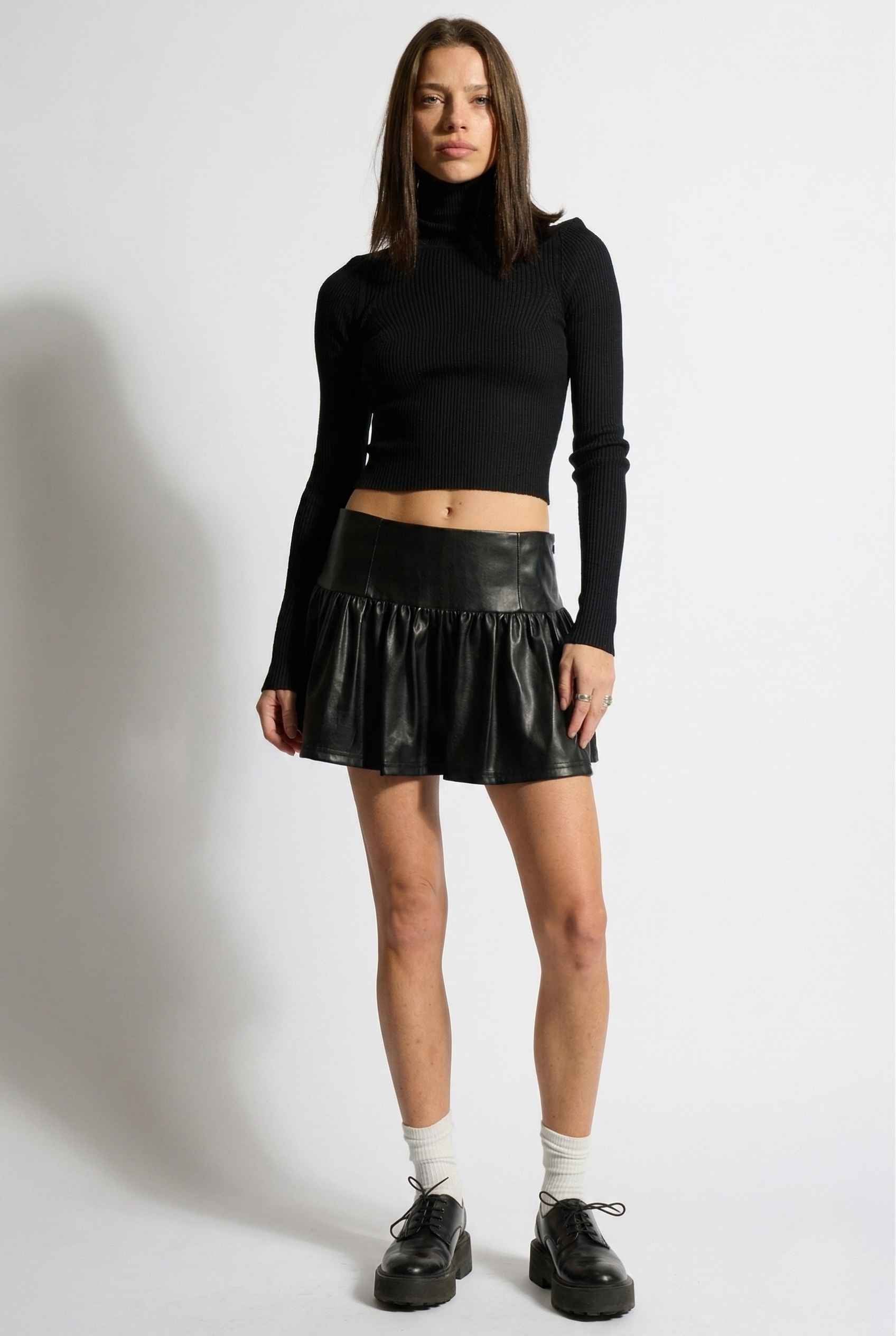 Woman wearing a black turtleneck and Nova Skirt Black Faux Leather Drop-Waist Ruffle Mini Skirt on a white background