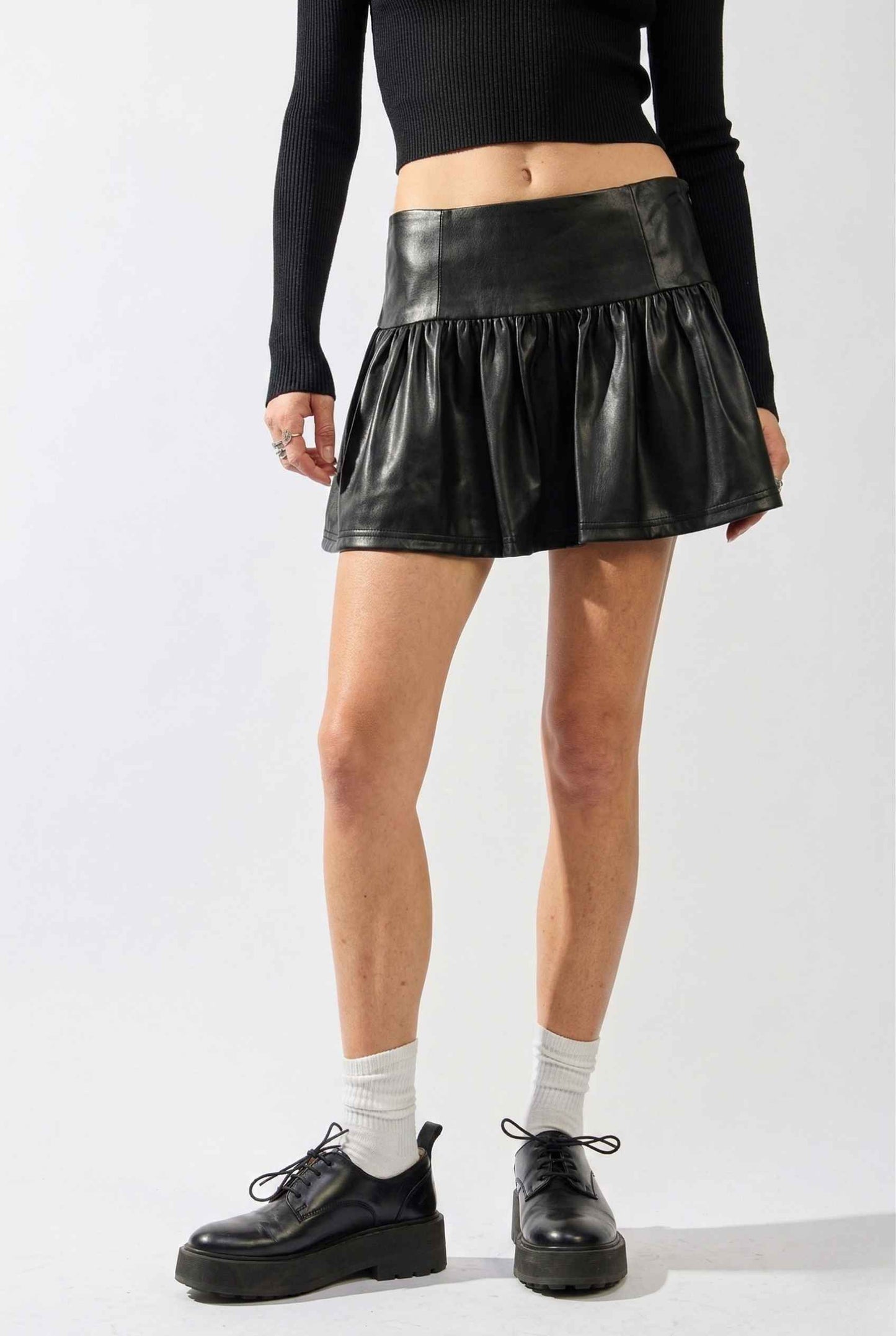 Woman wearing a Nova Skirt Black Faux Leather Drop-Waist Ruffle Mini Skirt with a plain background