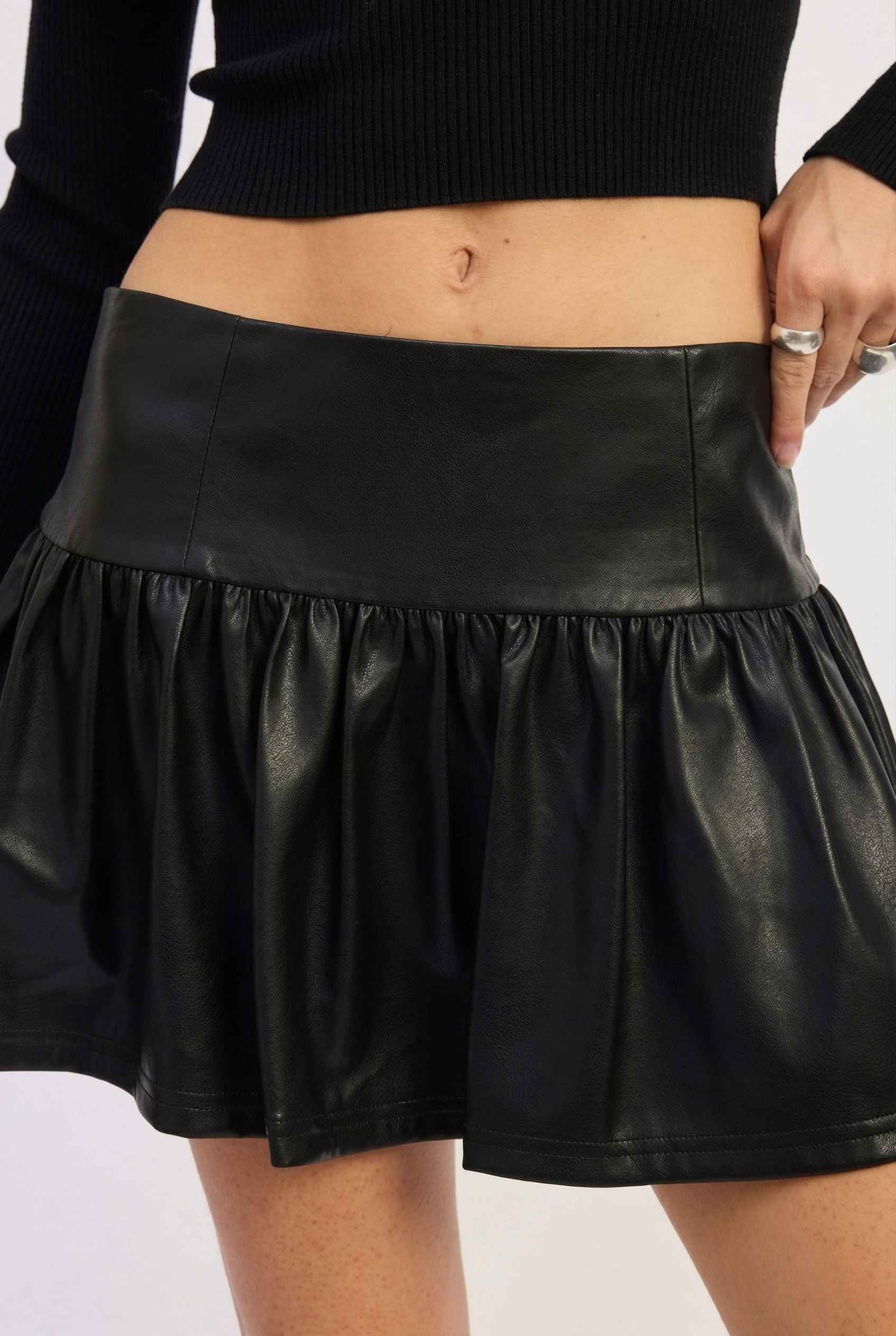 Nova Skirt Black Faux Leather Drop-Waist Ruffle Mini Skirt worn by a woman on a white background