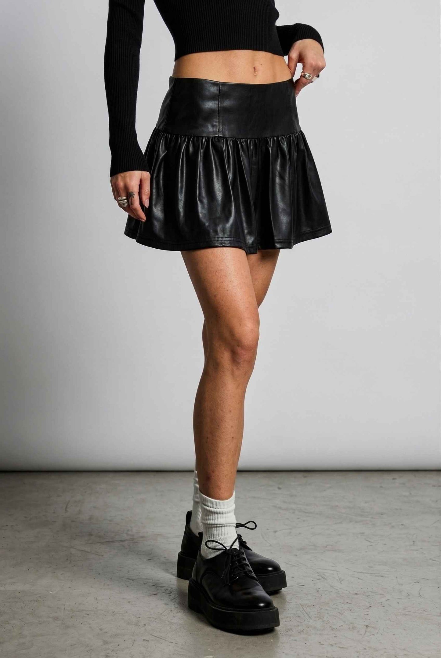 Woman wearing a Nova Skirt Black Faux Leather Drop-Waist Ruffle Mini Skirt with a plain background