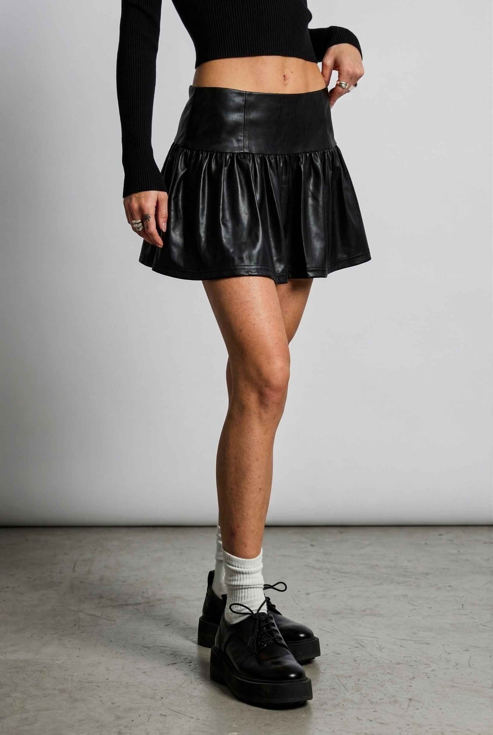 Woman wearing a Nova Skirt Black Faux Leather Drop-Waist Ruffle Mini Skirt with a plain background