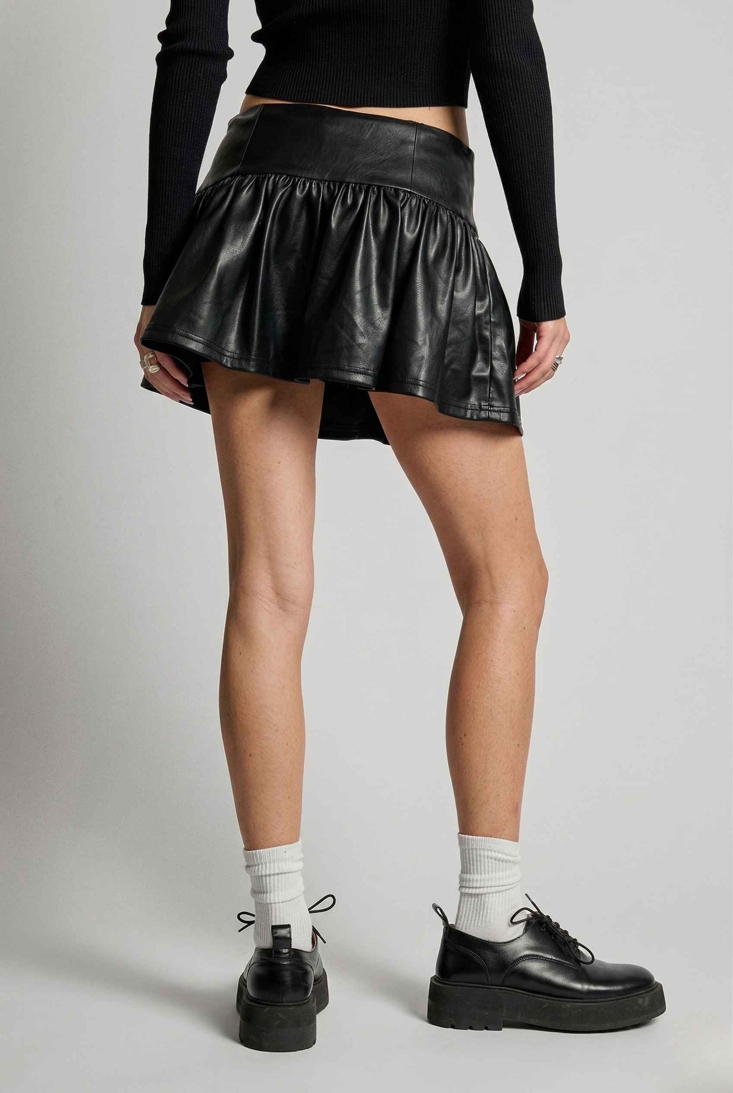 Woman wearing a Nova Skirt Black Faux Leather Drop-Waist Ruffle Mini Skirt with a plain background