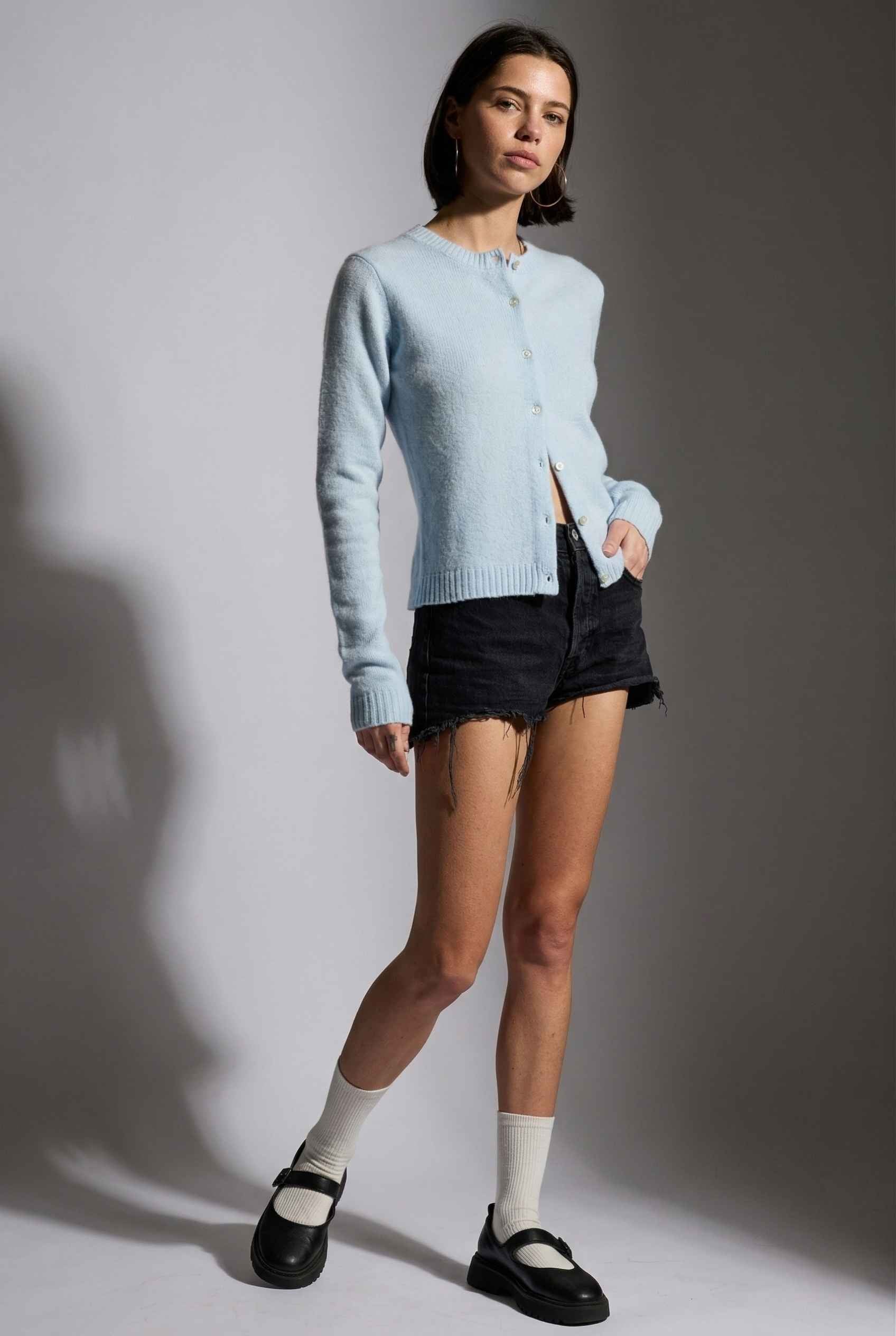 Model wearing Penelope Top in Blue Soft Wool-Blend Crew Neck Button-Up Cardigan with ripped jean shorts and mary janes