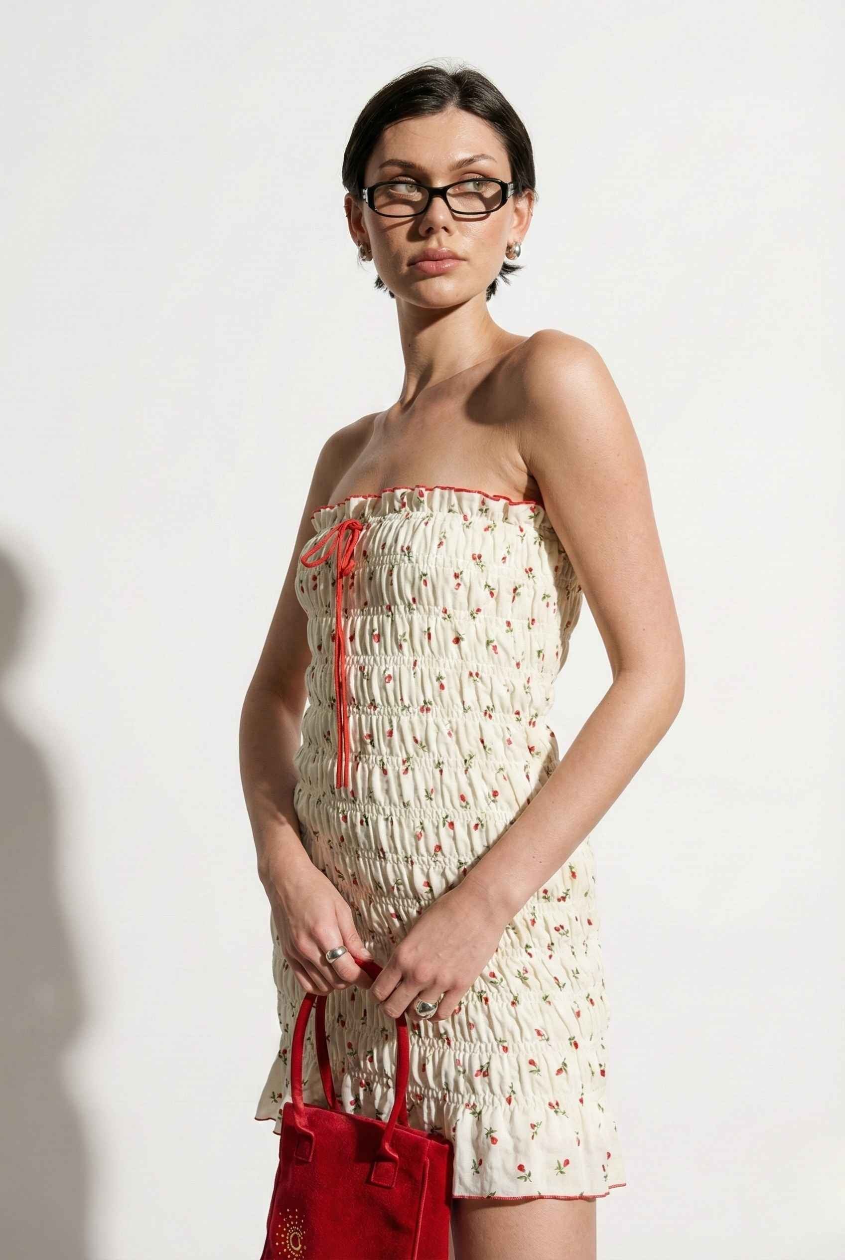 Woman wearing a Pippa Mini Cream Smocked Cherry Print Strapless Dress with red accents and holding a red handbag against a white background