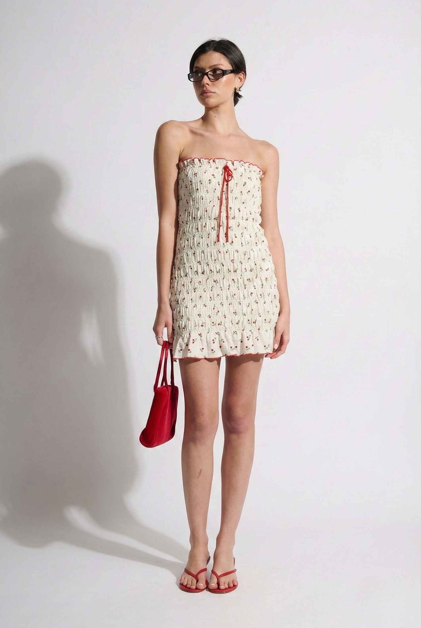 Woman wearing a Pippa Mini Cream Smocked Cherry Print Strapless Dress holding a red handbag against a white background