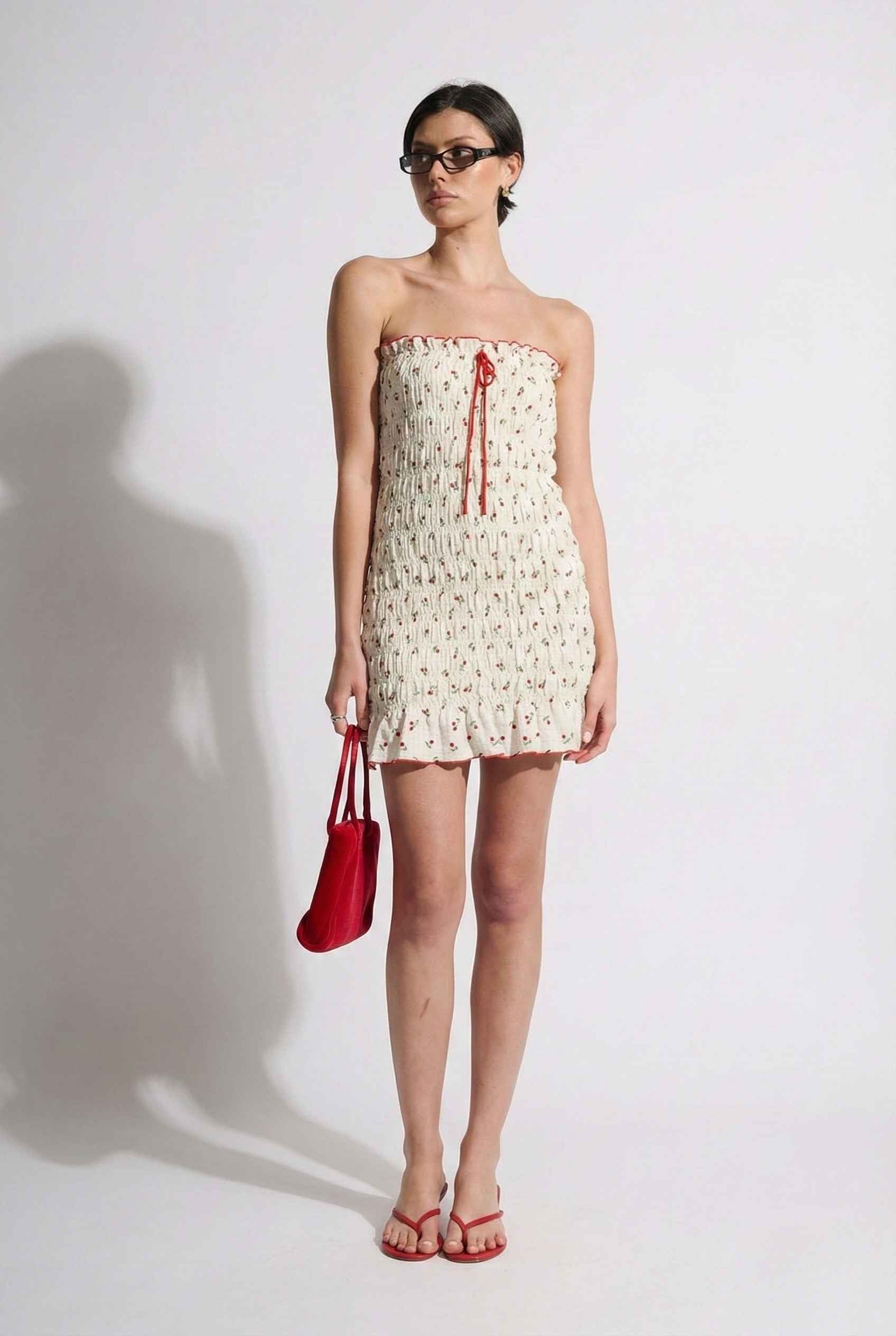 Woman wearing a Pippa Mini Cream Smocked Cherry Print Strapless Dress holding a red handbag against a white background