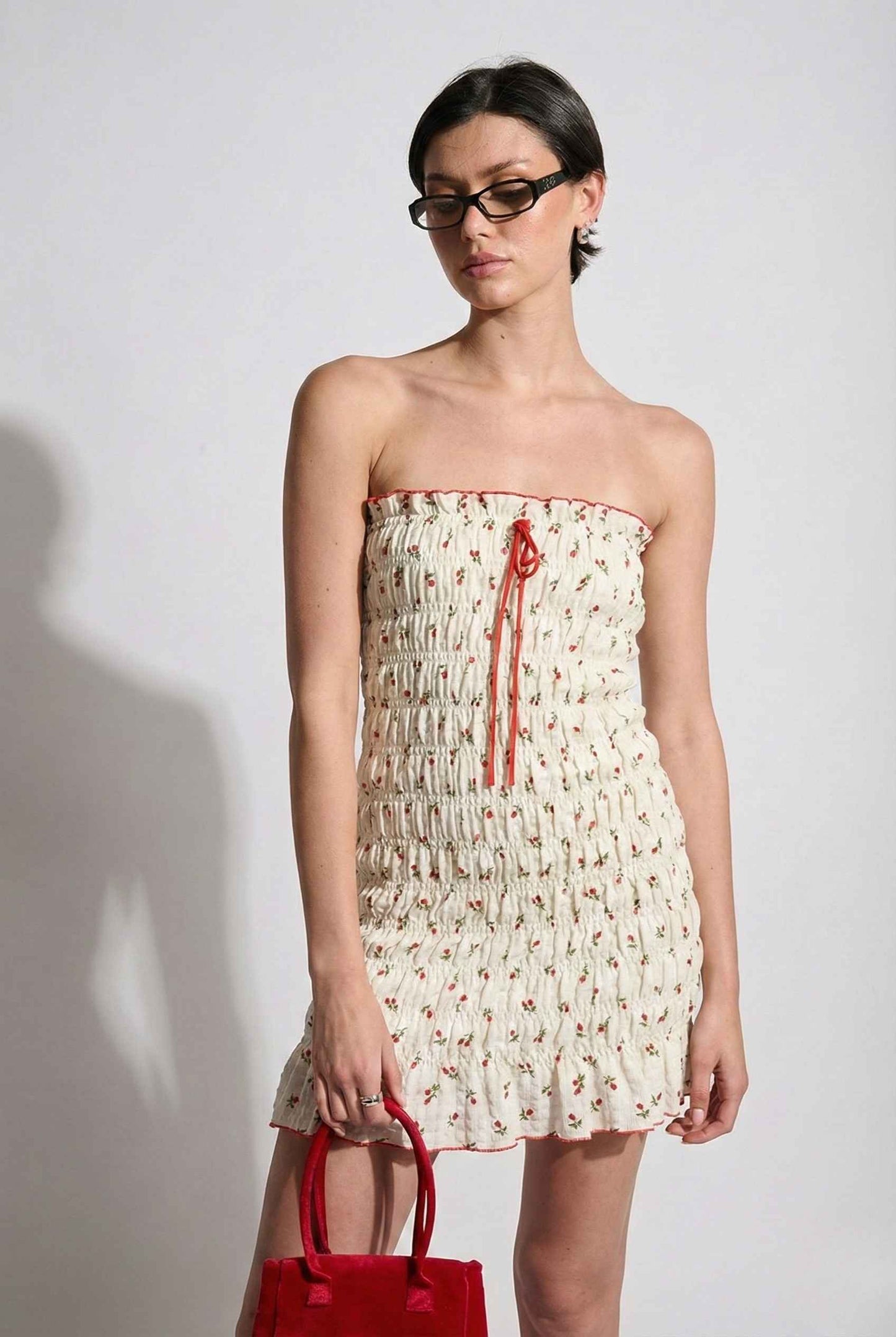Woman wearing a Pippa Mini Cream Smocked Cherry Print Strapless Dress holding a red handbag against a white background