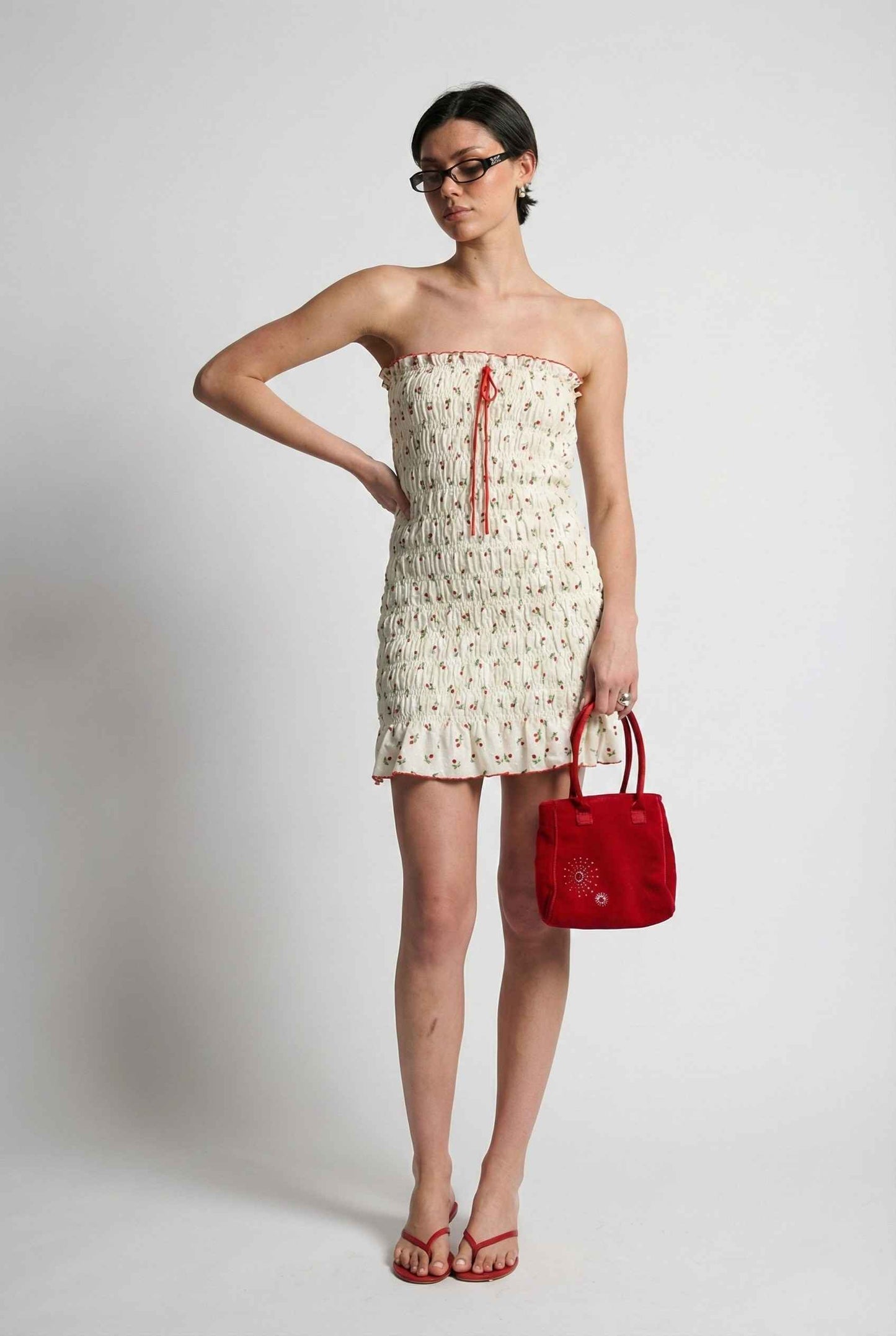Woman wearing a Pippa Mini Cream Smocked Cherry Print Strapless Dress with floral pattern and holding a red handbag against a plain background