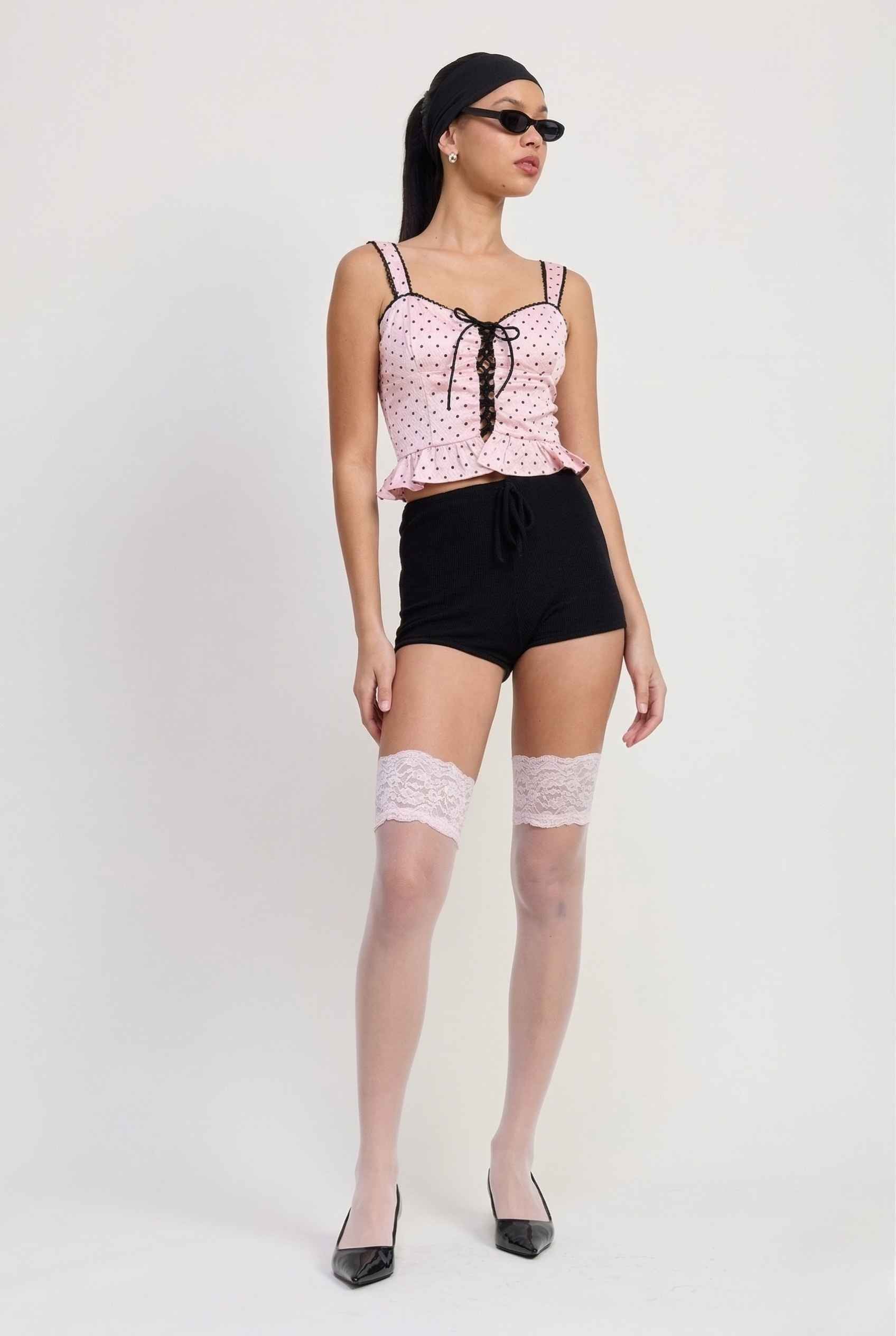 Woman wearing a Pippa Top Pink & Black Polka Dot Lace Trim Corset, black shorts, and white stockings on a white background