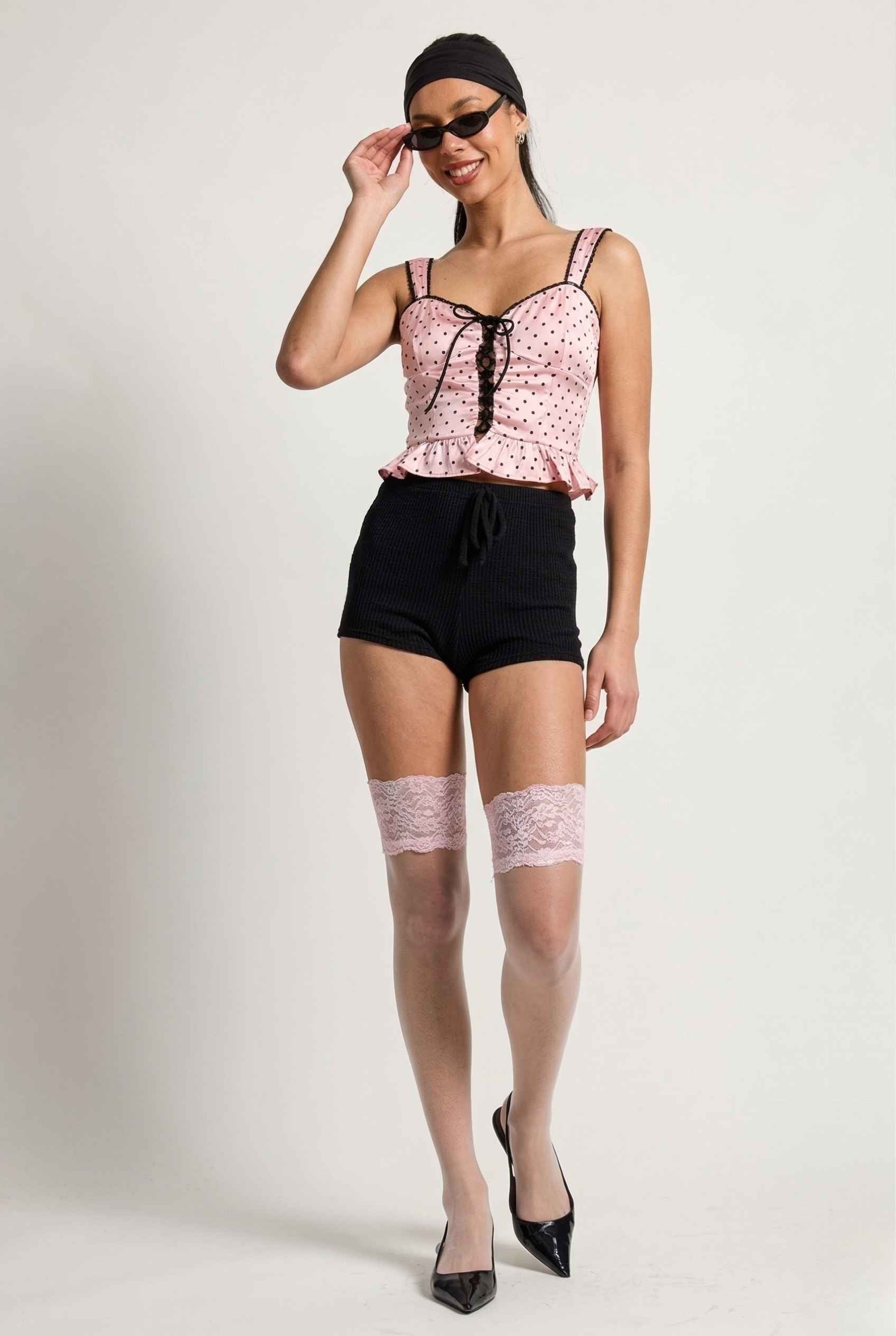 Woman wearing a Pippa Top Pink & Black Polka Dot Lace Trim Corset, black shorts, and sheer stockings with lace trim on a white background