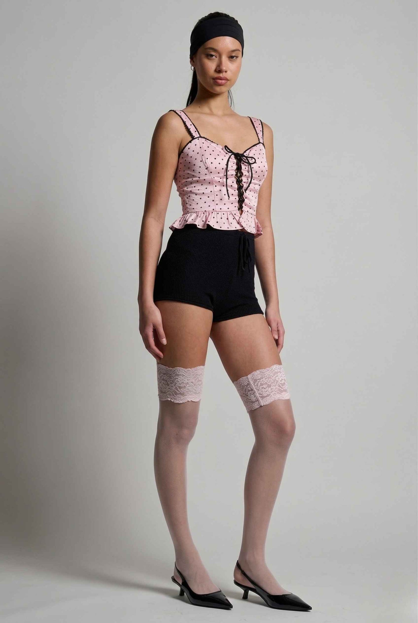 Woman wearing a Pippa Top Pink & Black Polka Dot Lace Trim Corset, black shorts, and lace knee-high socks against a plain background