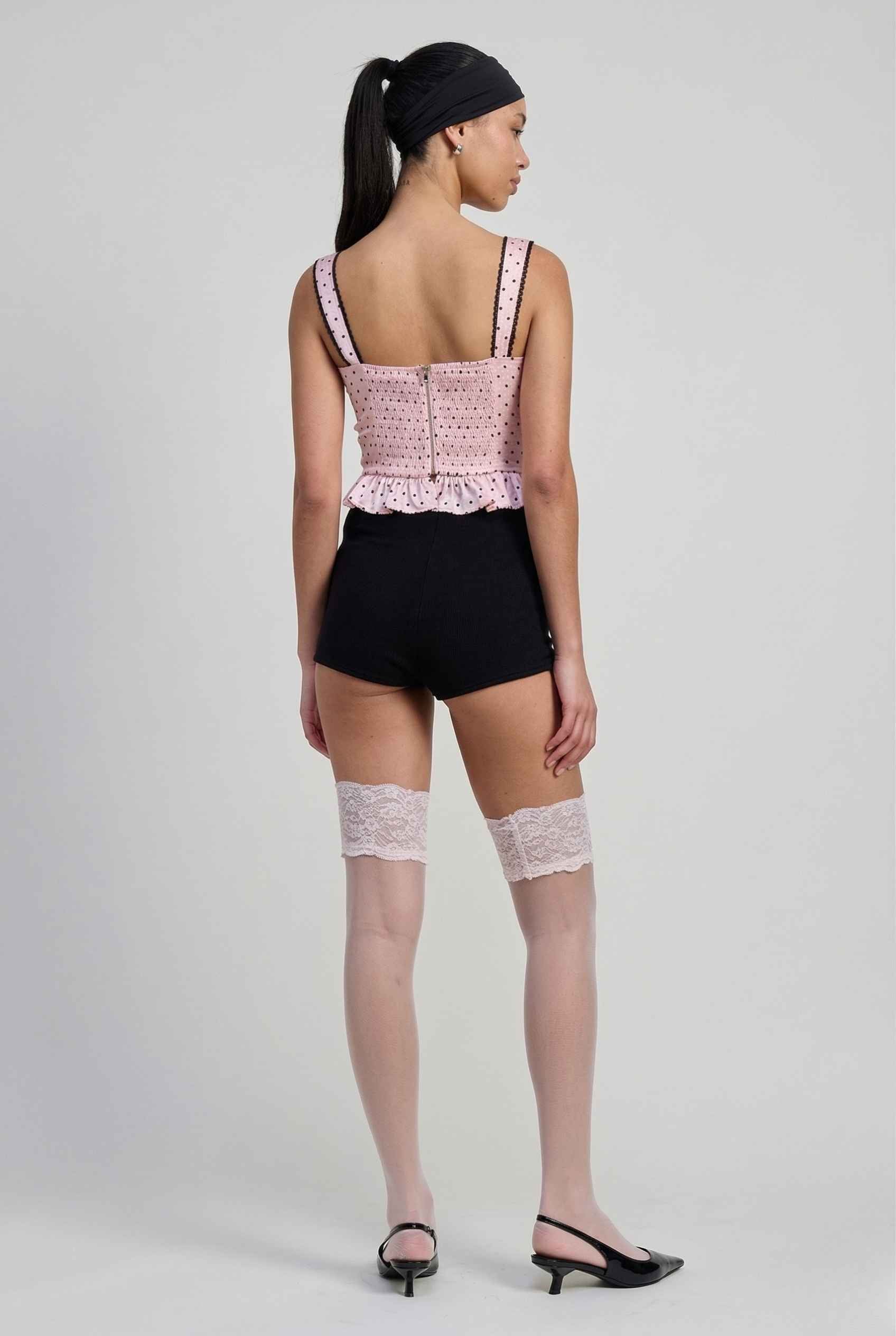 Woman wearing a Pippa Top Pink & Black Polka Dot Lace Trim Corset and black shorts with lace stockings on a plain background