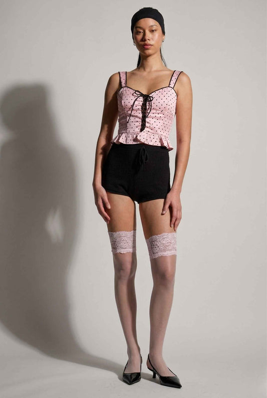 Woman wearing a Pippa Top Pink & Black Polka Dot Lace Trim Corset, black shorts, and sheer stockings with lace top on a plain background