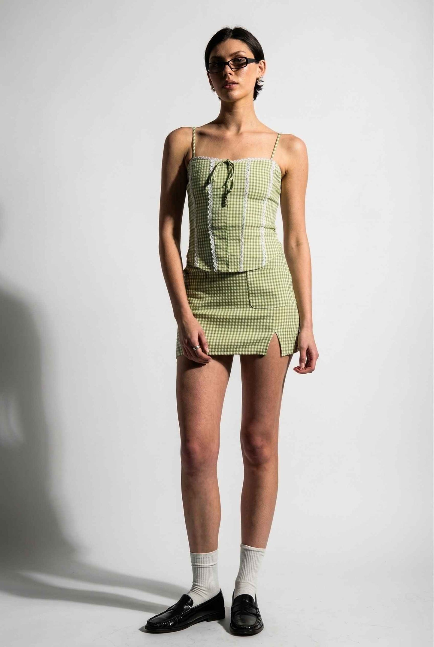 Model wearing Poppy Skirt in Sage Green Gingham Mini Skirt with Pockets paired matching corset tank top and shiny loafers with tall socks