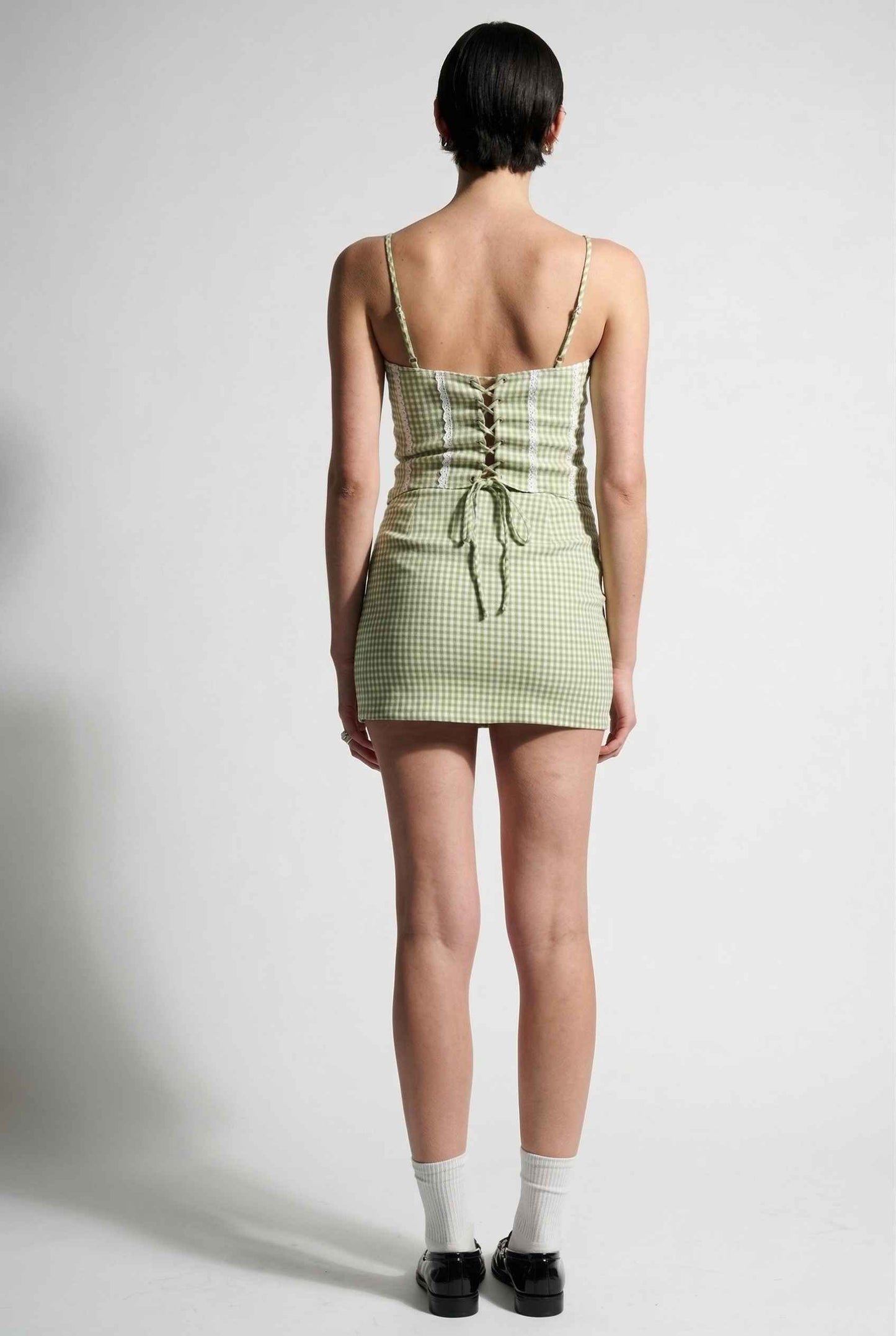 Model wearing Poppy Skirt in Sage Green Gingham Mini Skirt with Pockets paired matching corset tank top and shiny loafers with tall socks
